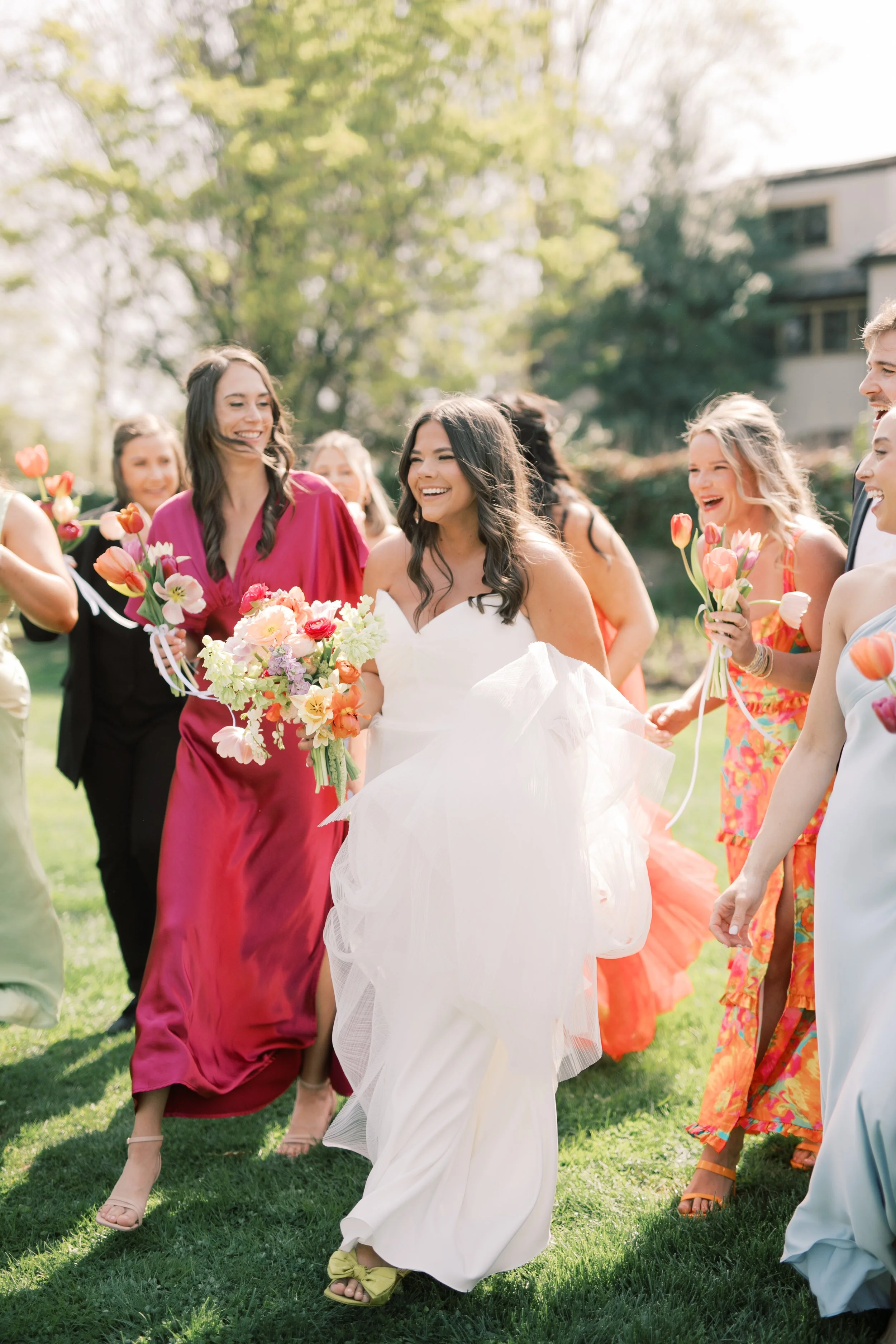 A bride in a white dress and yellow shoes walking through a group of smiling friends holding flowers outdoors on a sunny day.