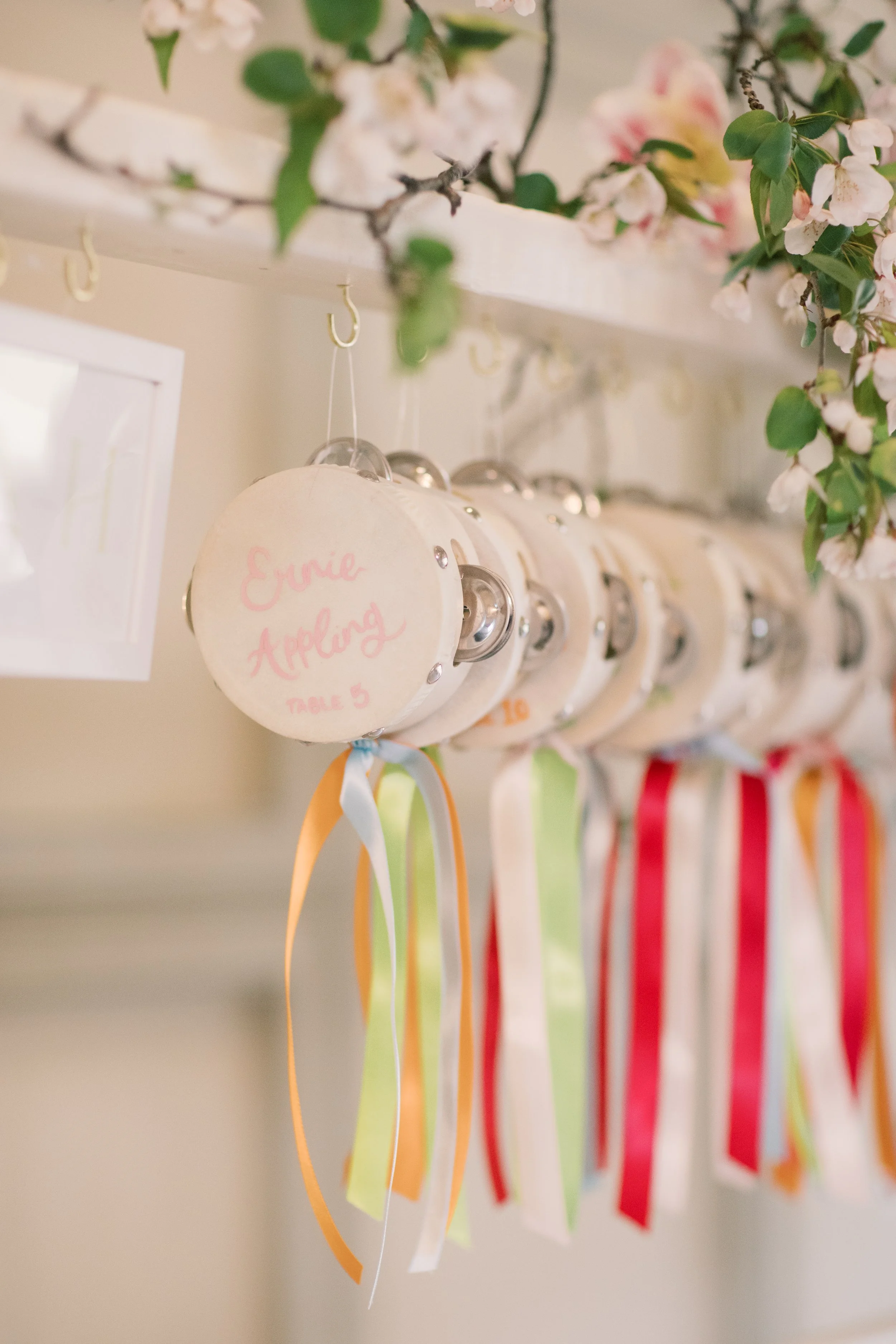 Decorative tambourines with colorful ribbons hanging from a white shelf decorated with pink and white flowers.