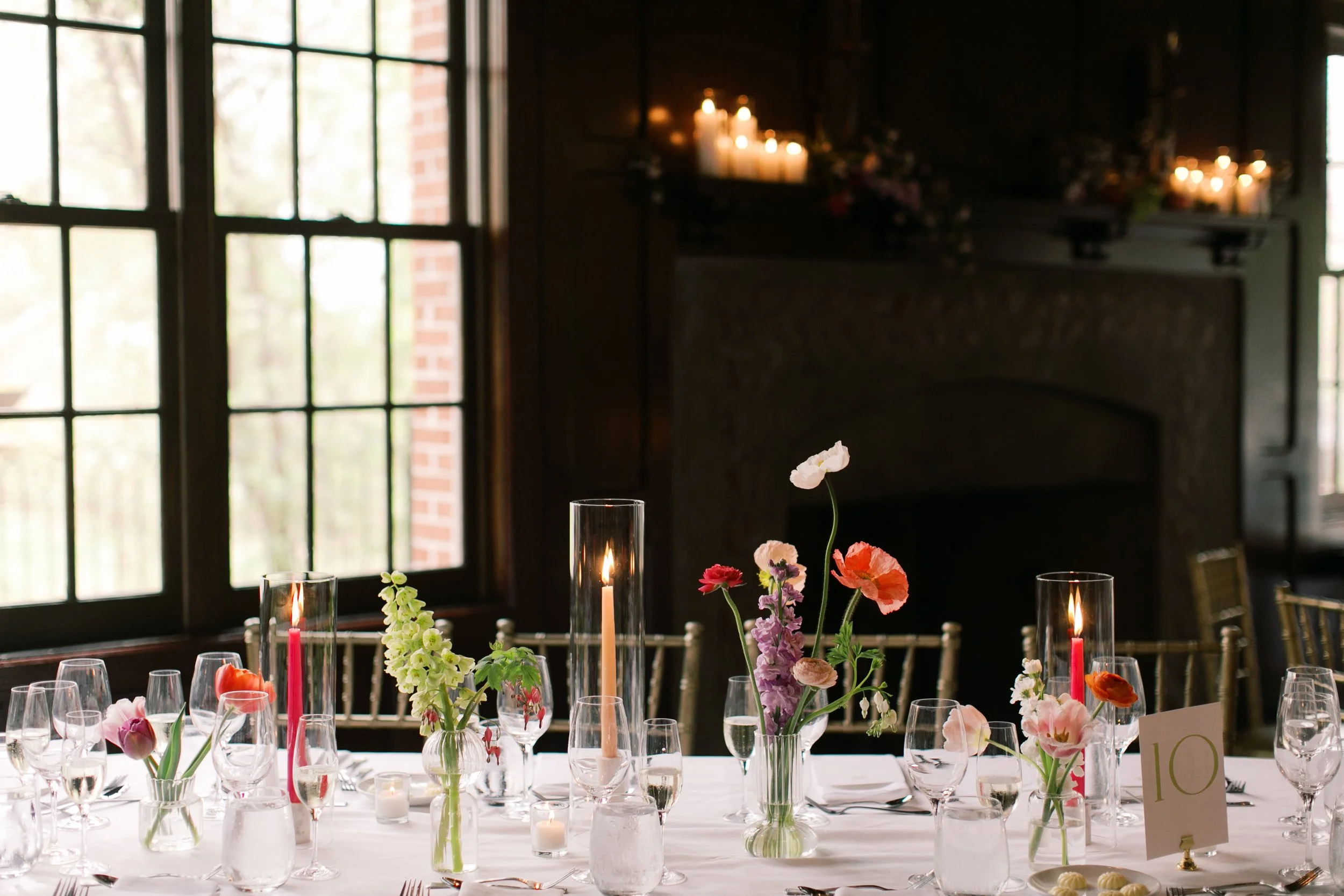 Elegant dining table decorated with pink and purple flowers in glass vases, surrounded by candles and glasses, in a dimly lit room with large windows and dark walls.
