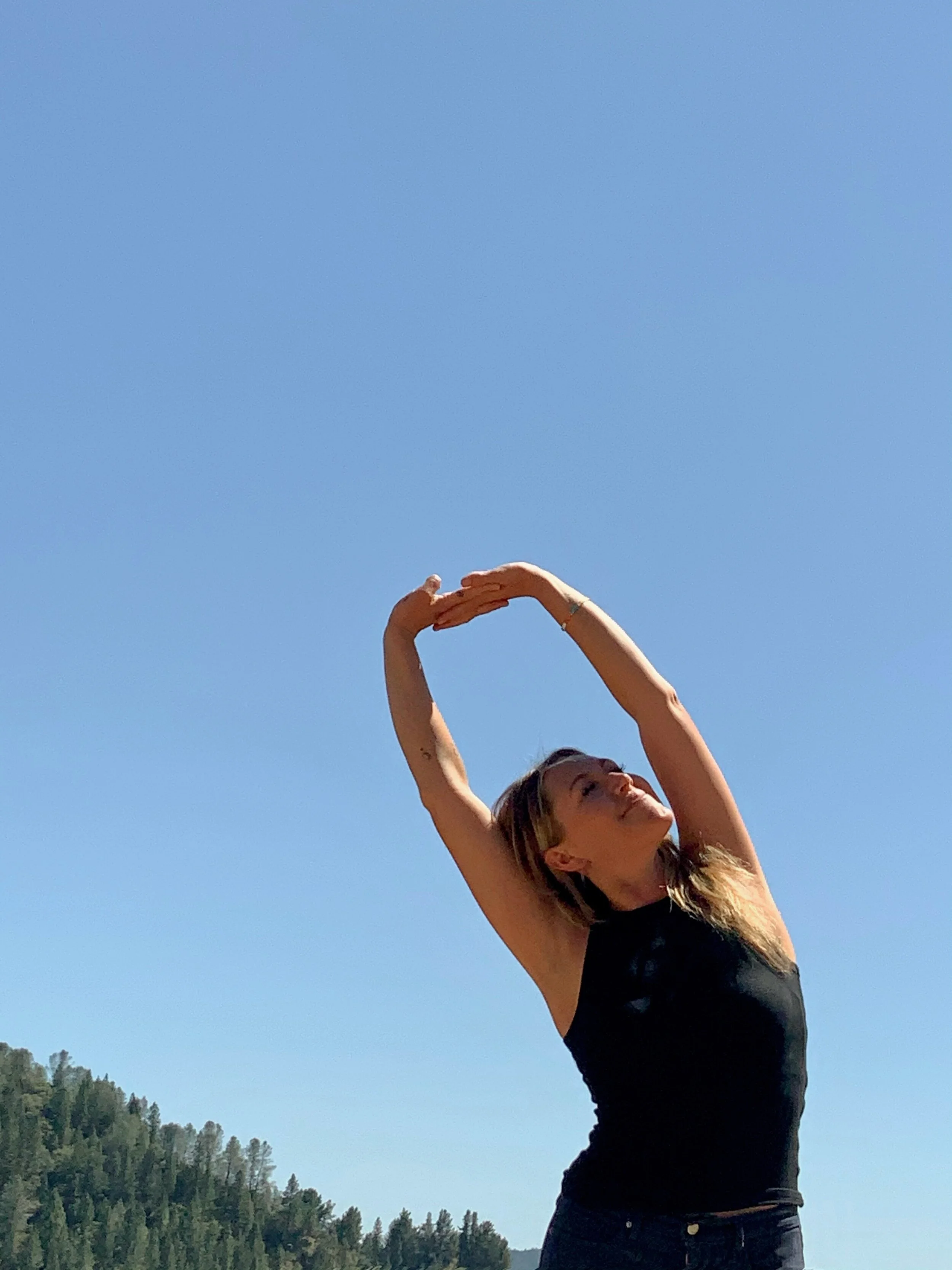 A woman with long blonde hair wearing a black sleeveless top and jeans standing outdoors with arms raised, forming a heart shape with her hands against a bright blue sky with some trees and hills in the background.