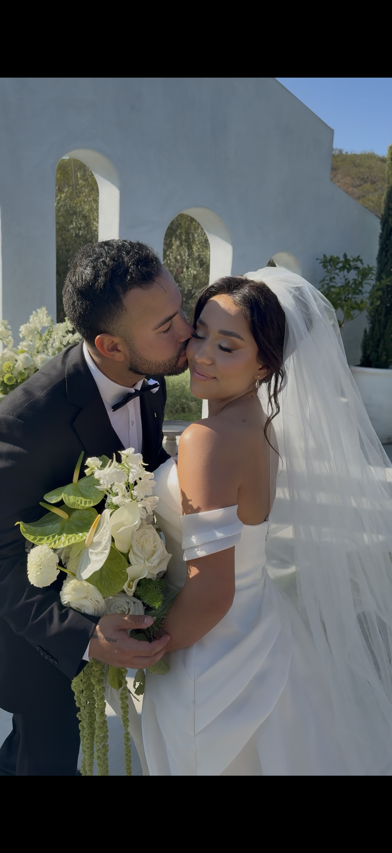 A bride and groom share a kiss during their wedding ceremony outdoors, with the groom in a black tuxedo and the bride in a white wedding gown holding a bouquet of white and green flowers.