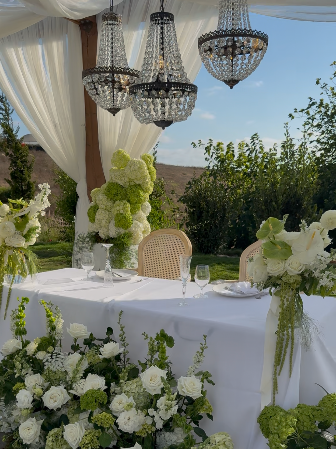 An elegant outdoor table set for a celebration with white flowers and greenery, under a canopy with hanging chandeliers, with a scenic landscape in the background.