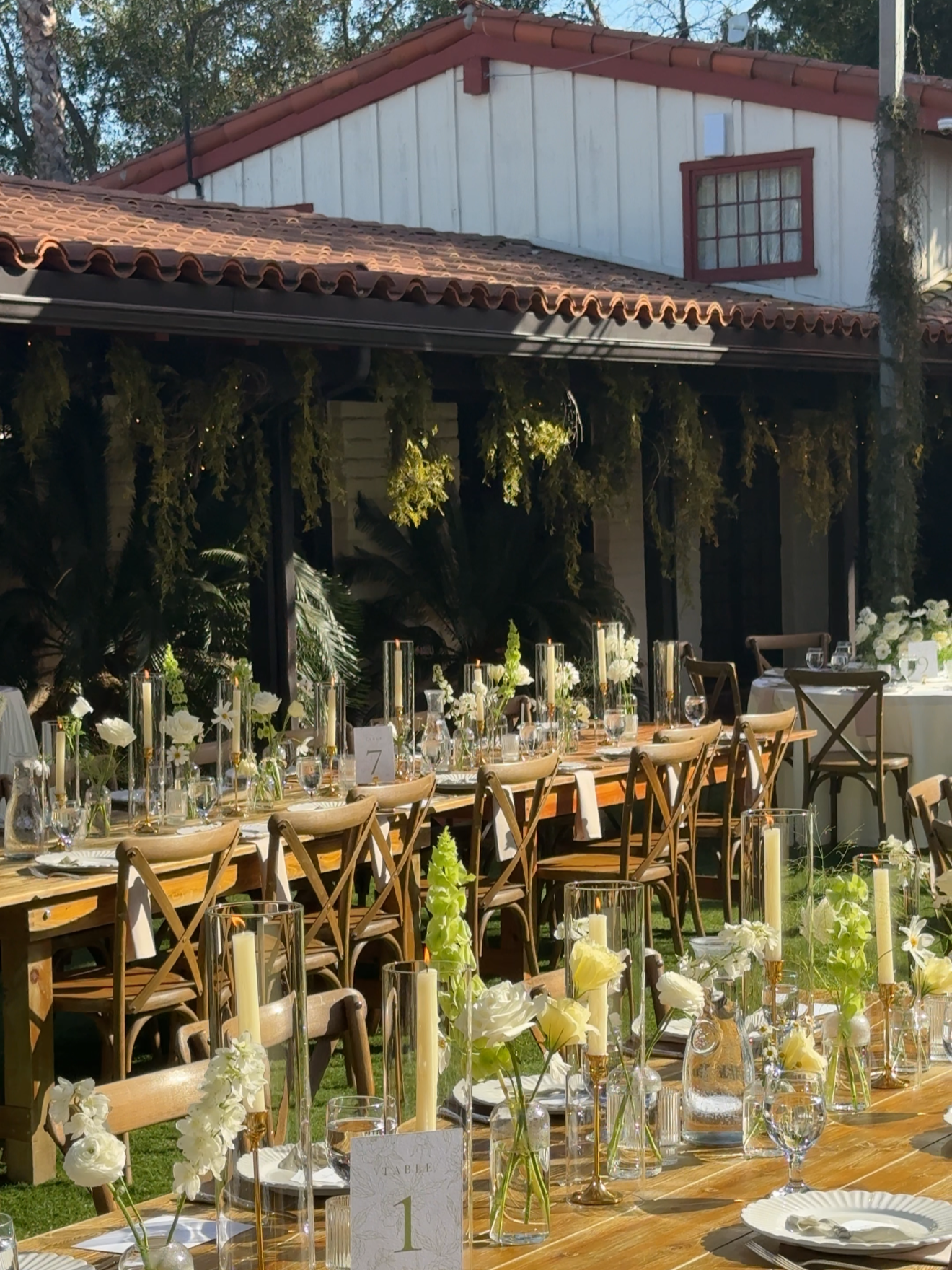 Wedding reception setup with decorated tables, flowers, candles, and white tableware outdoors in a garden with a house in the background.
