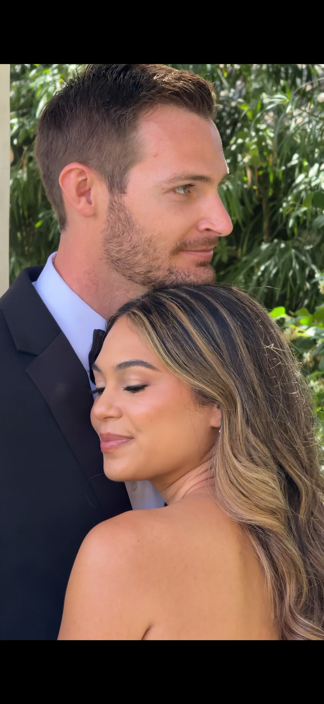 A man in a tuxedo and a woman with long hair are hugging outdoors with green foliage in the background, both with content expressions.