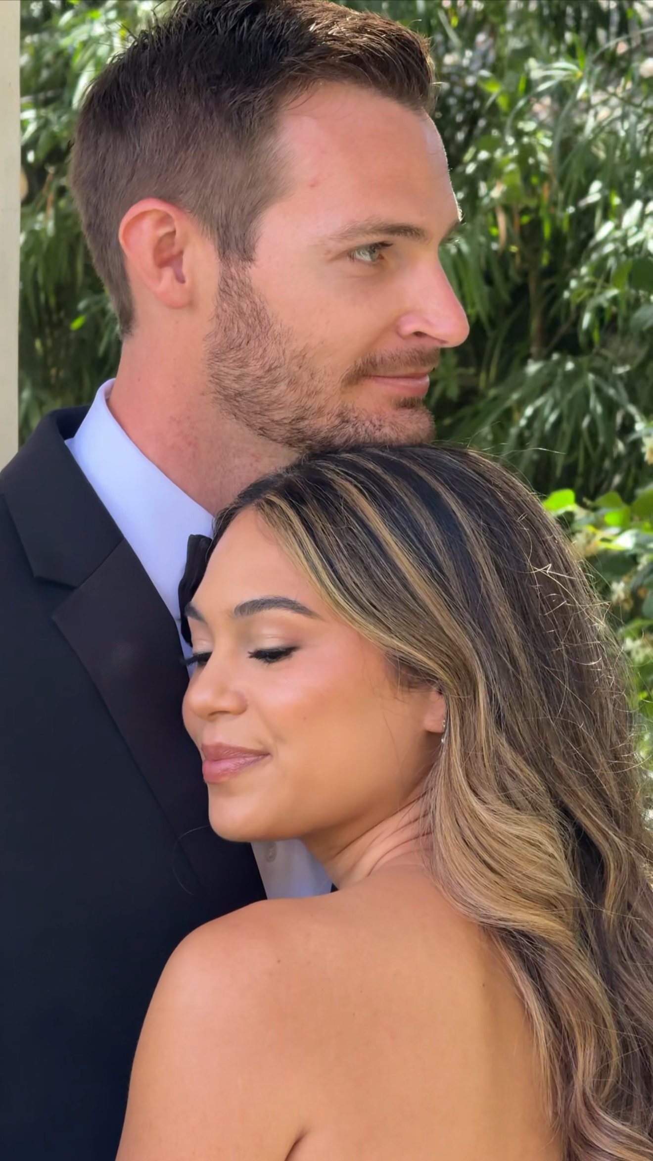 A man in a tuxedo and a woman with long hair in an embrace, with greenery in the background.
