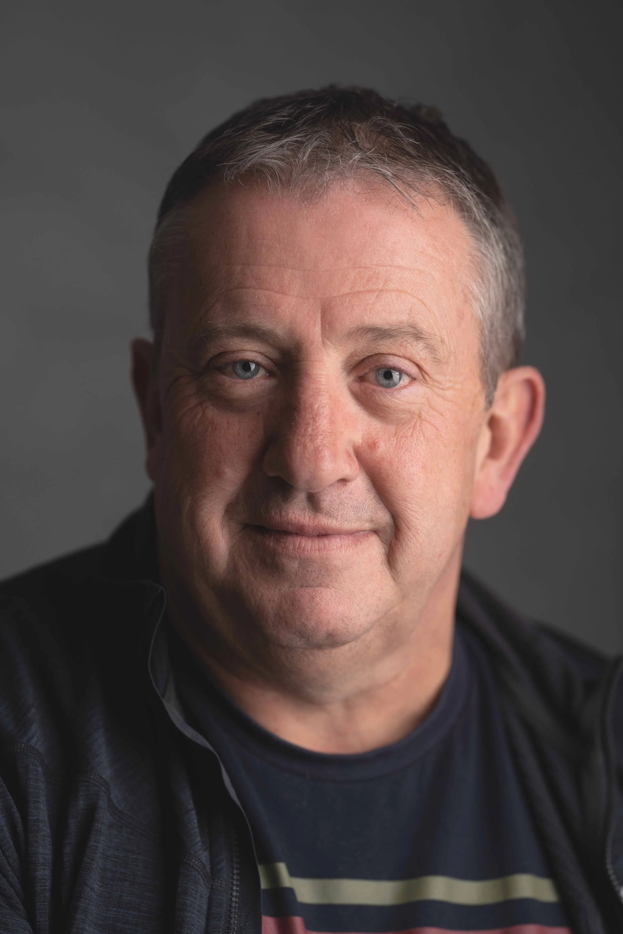 Close-up portrait of a middle-aged man with short gray hair, blue eyes, and light skin, smiling slightly, wearing a dark jacket and a T-shirt with horizontal stripes, against a dark gray background.