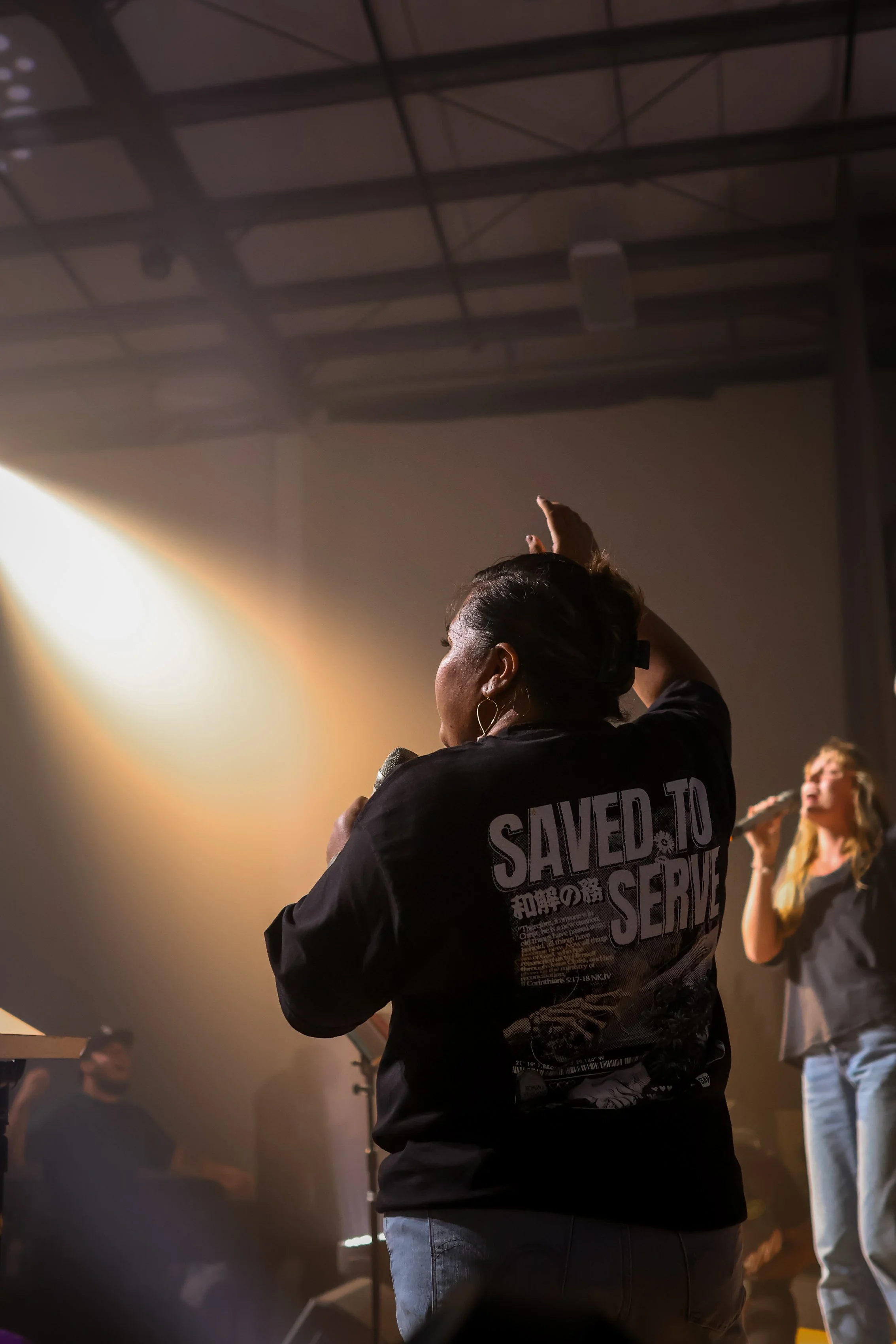 A woman with dark hair pulled back, holding a microphone, raising her other hand during a live performance or worship event inside a dimly lit industrial-style space, with a soft spotlight shining on her.