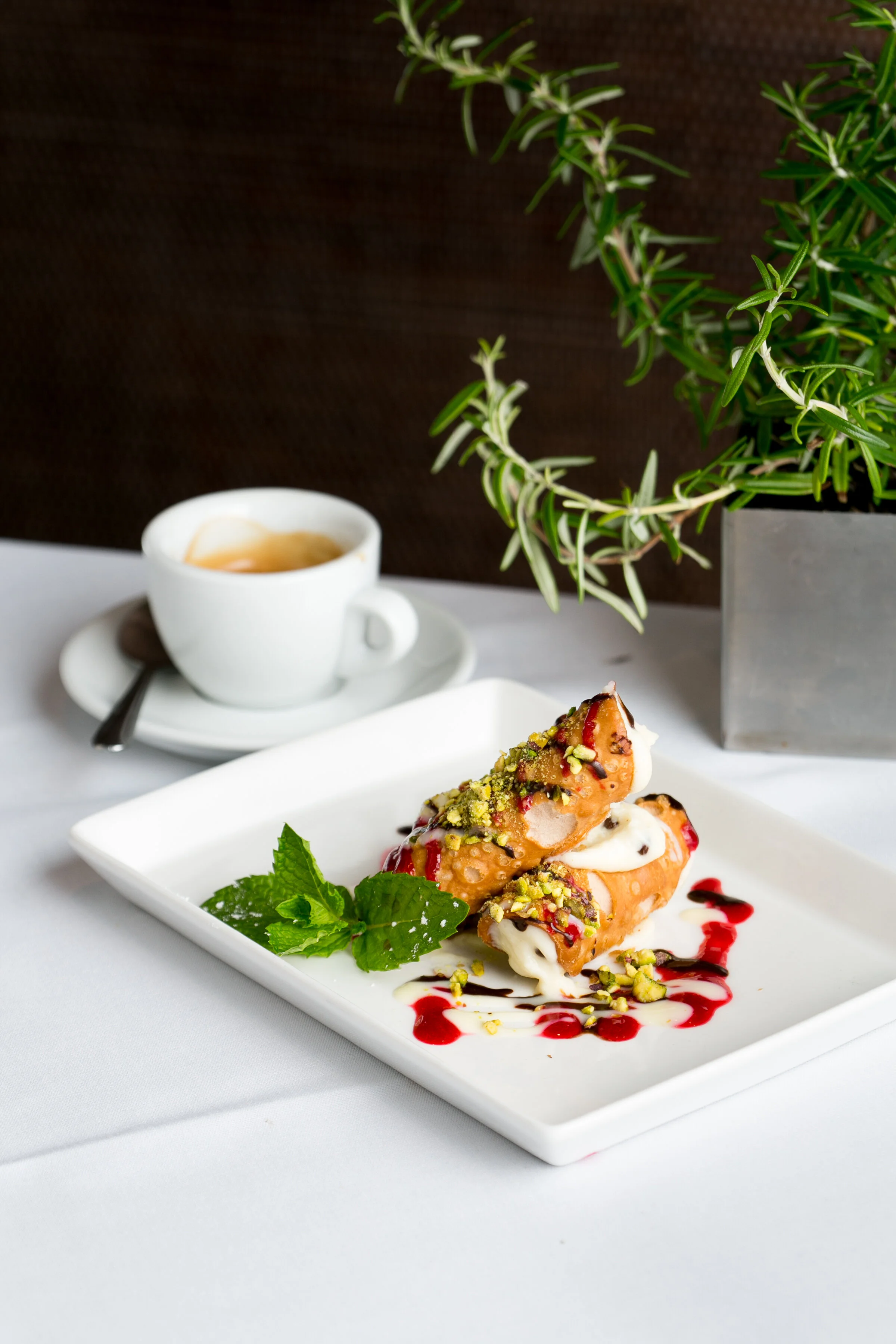 Two rolled ice cream crepes topped with pistachios, served with mint leaves on a white rectangular plate, with a cup of coffee in the background, on a white table with a potted plant nearby.