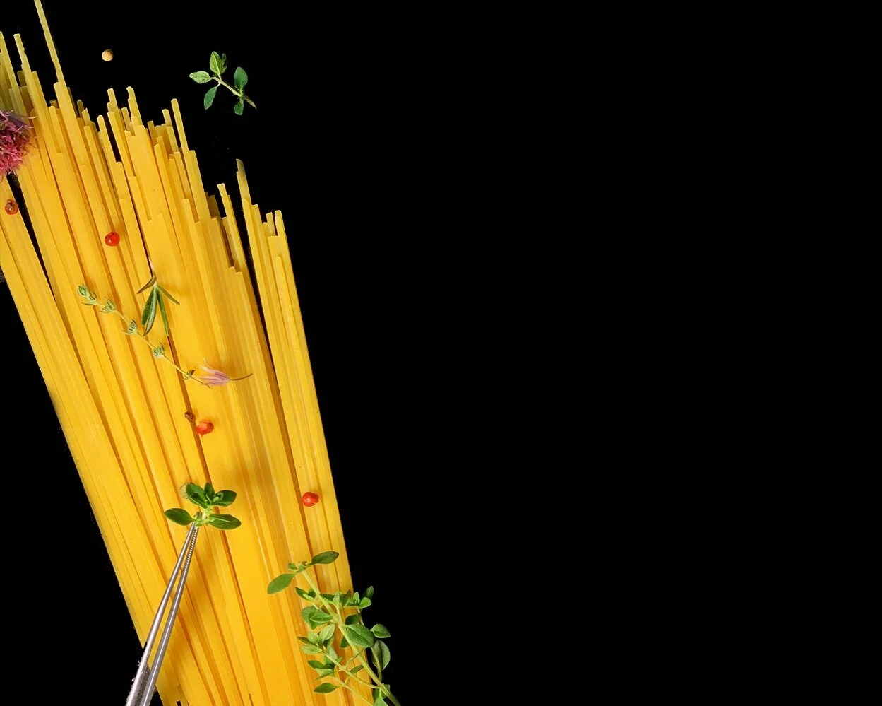 Uncooked spaghetti noodles with herbs and peppercorns on a black background.