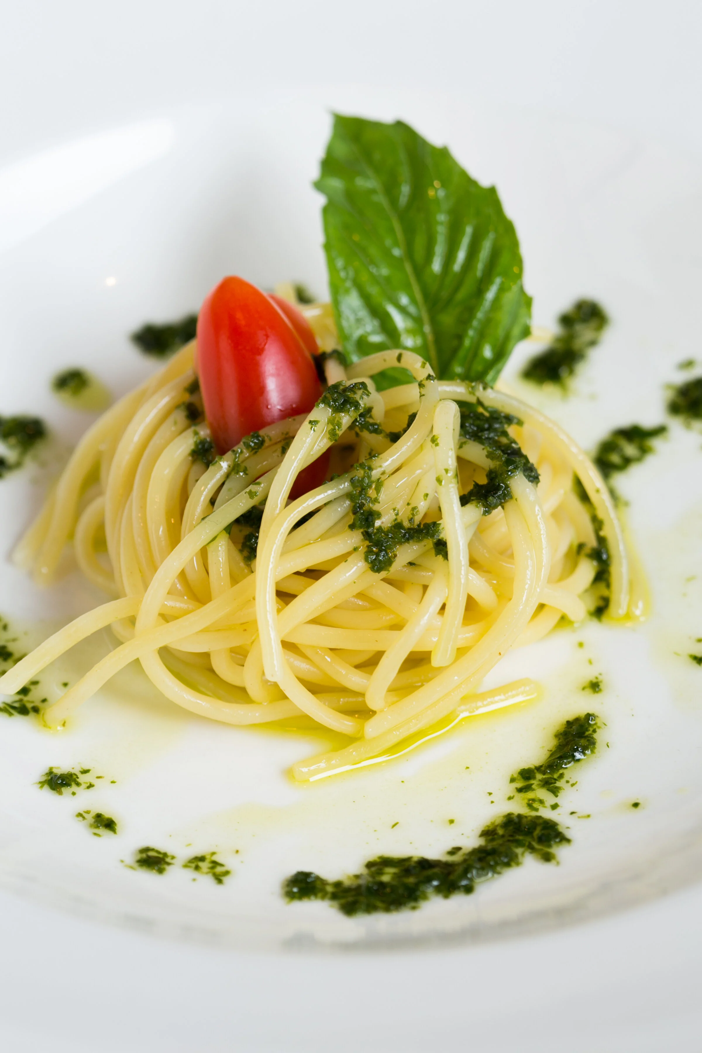 A plate of spaghetti with basil pesto, a cherry tomato, and a basil leaf.