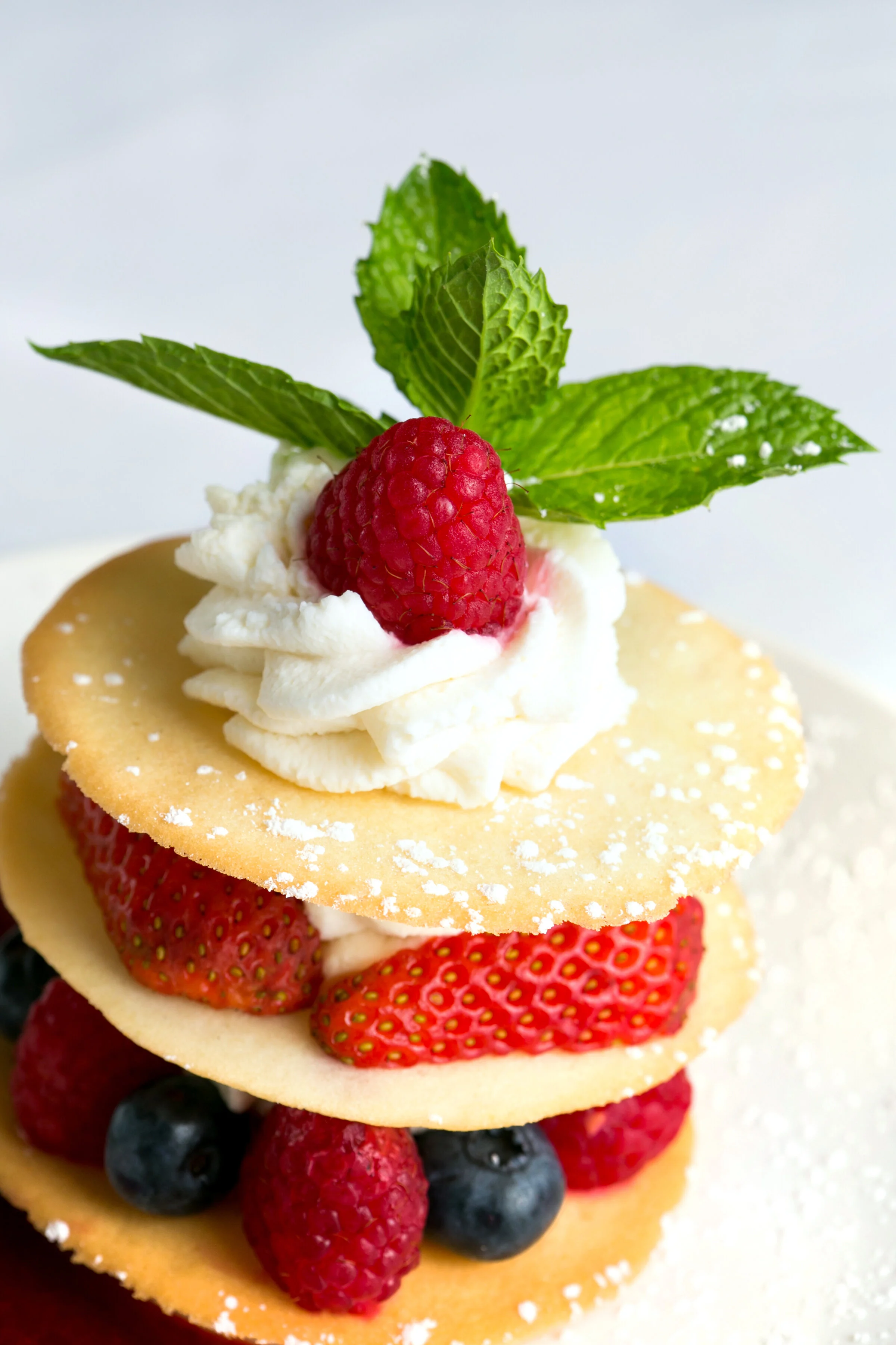 A layered dessert with fresh strawberries, blueberries, raspberry, and a raspberry on top, garnished with mint leaves and whipped cream, served between thin, crispy layers.