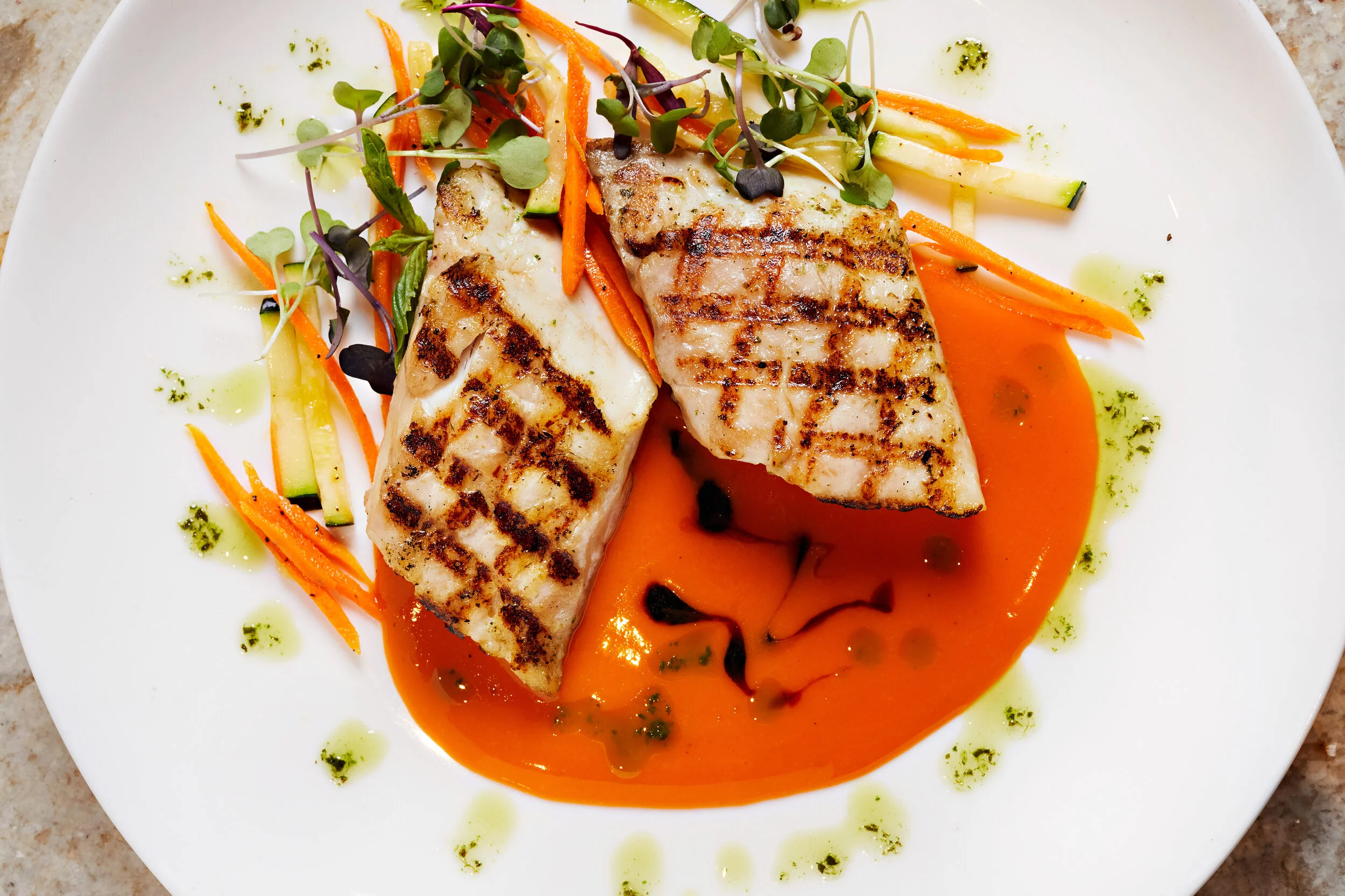 Grilled fish fillets on a white plate with orange sauce, garnished with microgreens, shredded carrots, cucumber strips, and drizzled with green and dark sauces.