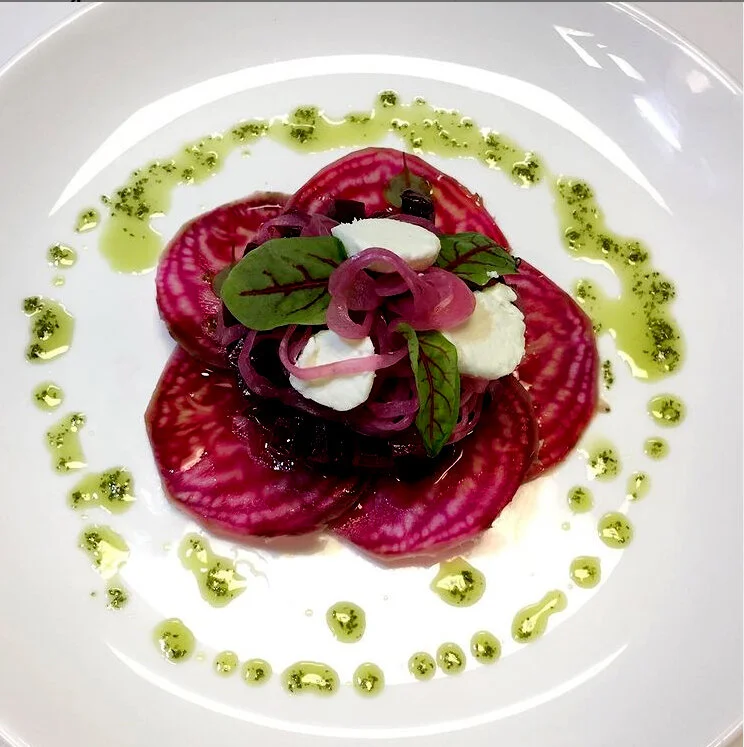 Beet salad with goat cheese, pickled onions, greens, and drizzled green dressing on a white plate.