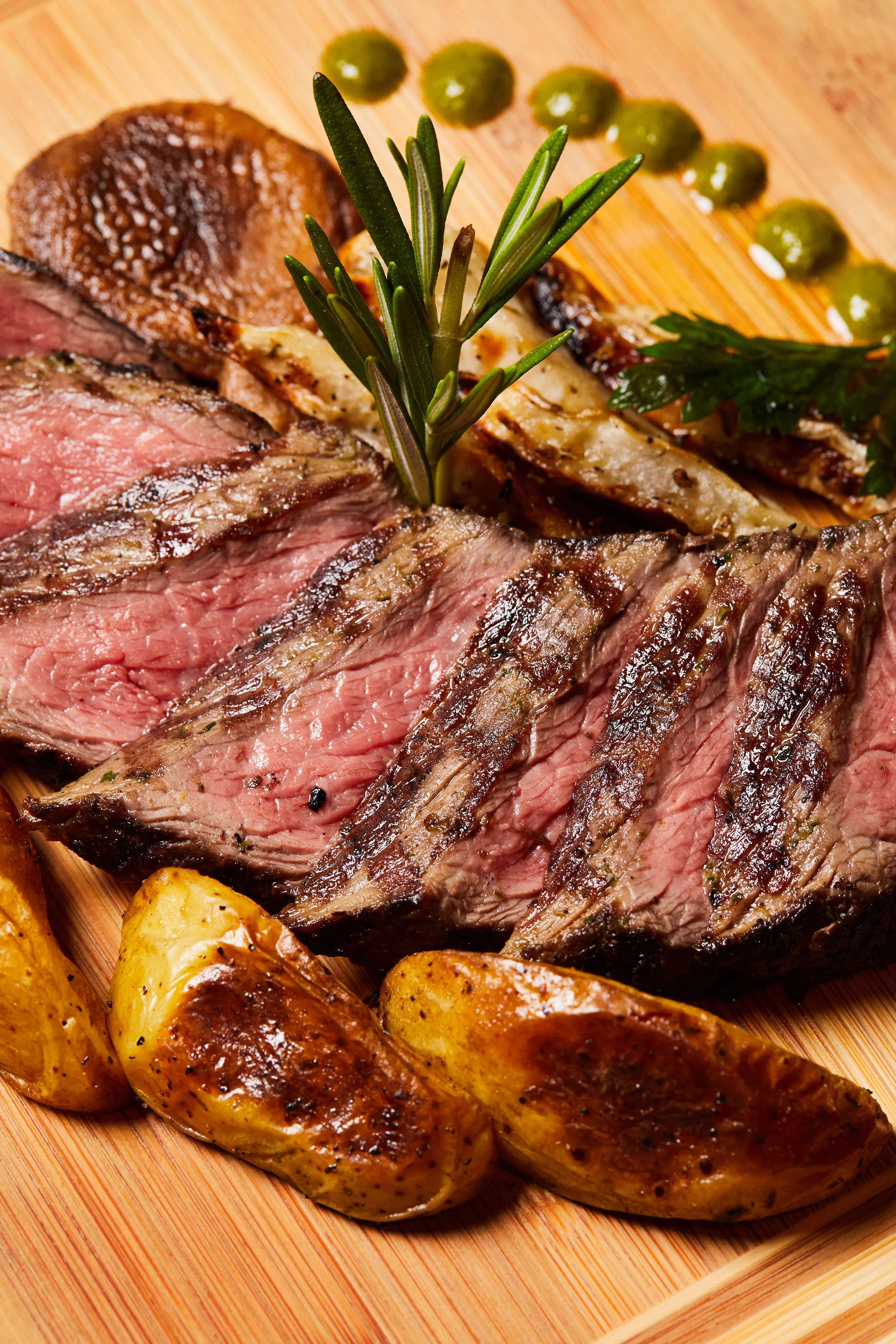 Sliced grilled steak topped with rosemary, accompanied by roasted potatoes and green sauce on a wooden cutting board.