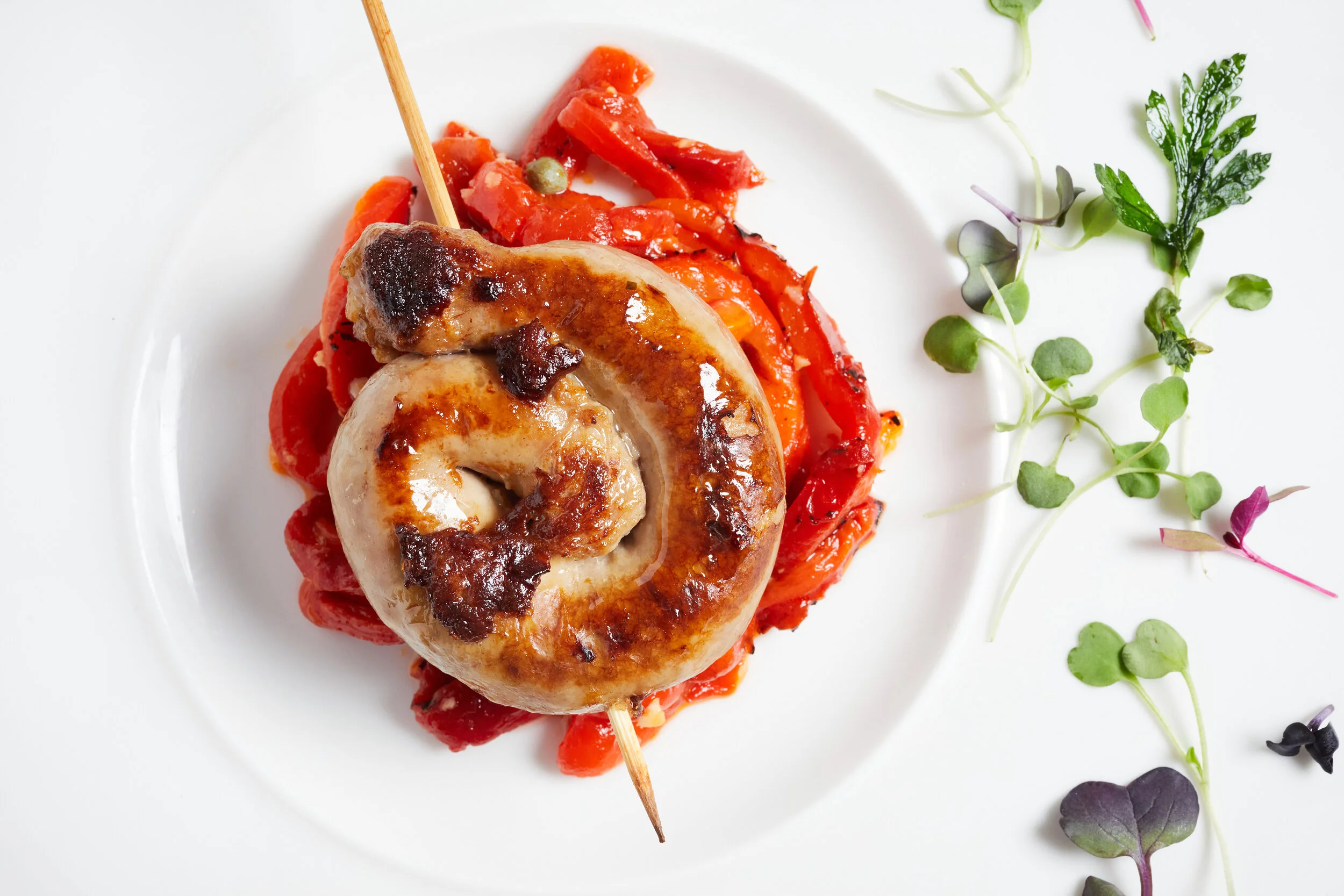A grilled chicken drumstick with a wooden skewer, placed on a bed of grilled red bell peppers, served on a white plate. There are microgreens on the side of the plate.