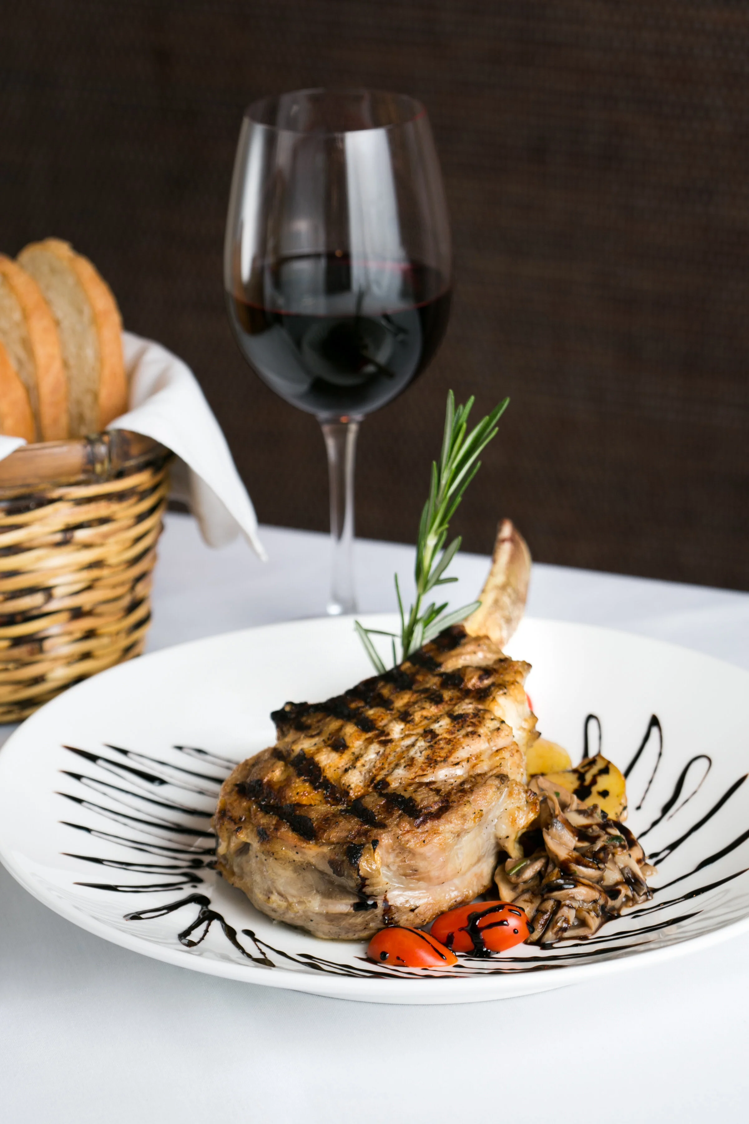 Grilled veal chop on a white plate with cherry tomatoes, mushroom sauté, and a sprig of rosemary, with a glass of red wine and bread in a basket in the background.