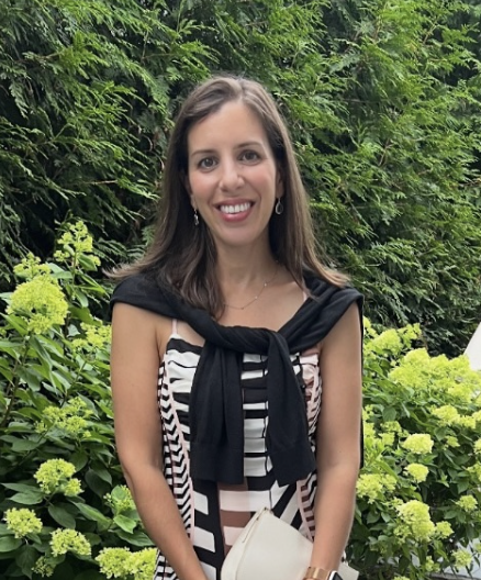 A woman with long brown hair smiling outdoors in front of green bushes with yellow flowers, wearing a black and white striped dress and a black sweater tied around her shoulders.