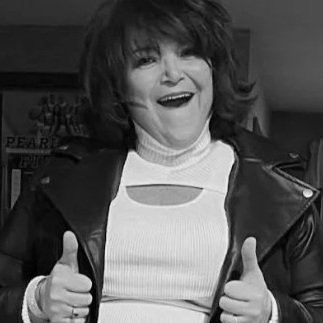 Black and white photo of a smiling woman with voluminous hair, wearing a white top and a dark jacket, giving two thumbs up.