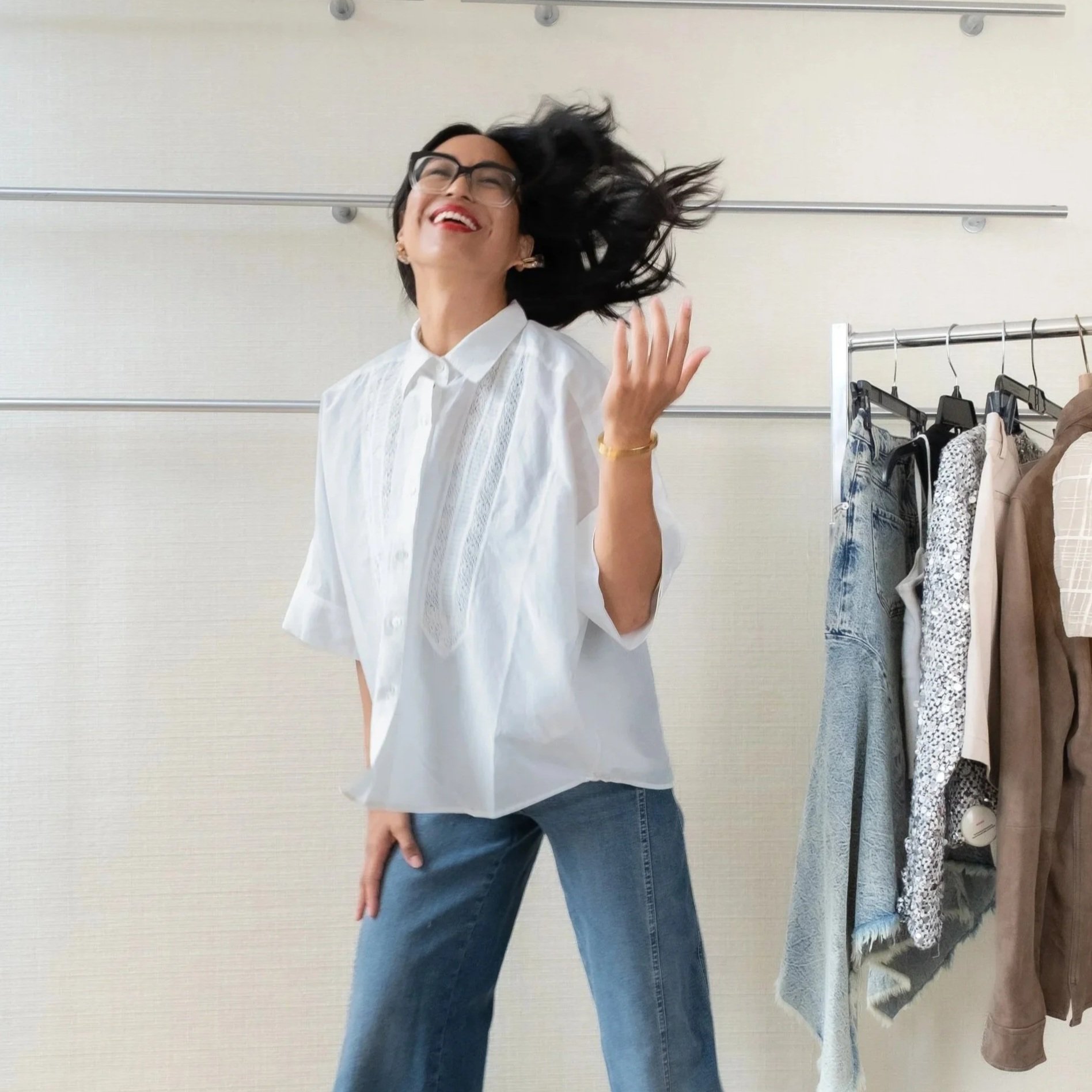 A woman with glasses, wearing a white blouse and blue jeans, is smiling and flipping her hair in a room with a clothing rack that has various garments hanging on it.