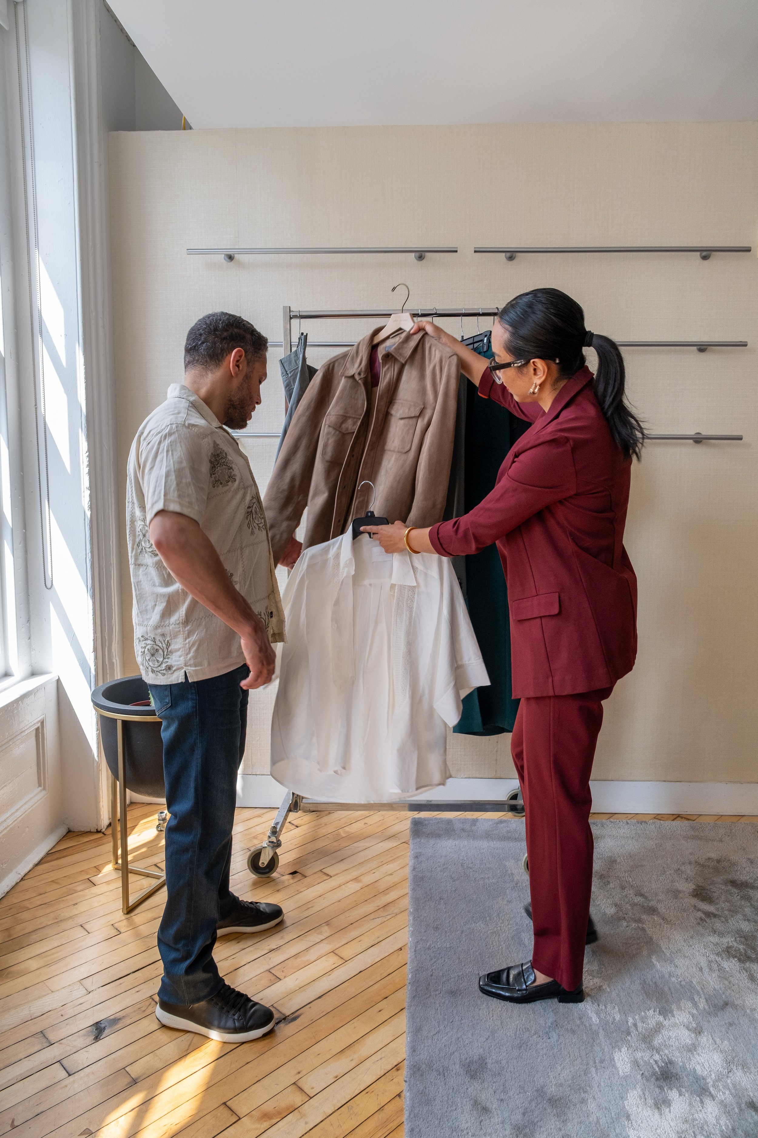 Two people looking at clothes on a clothing rack indoors, with sunlight coming through a window.