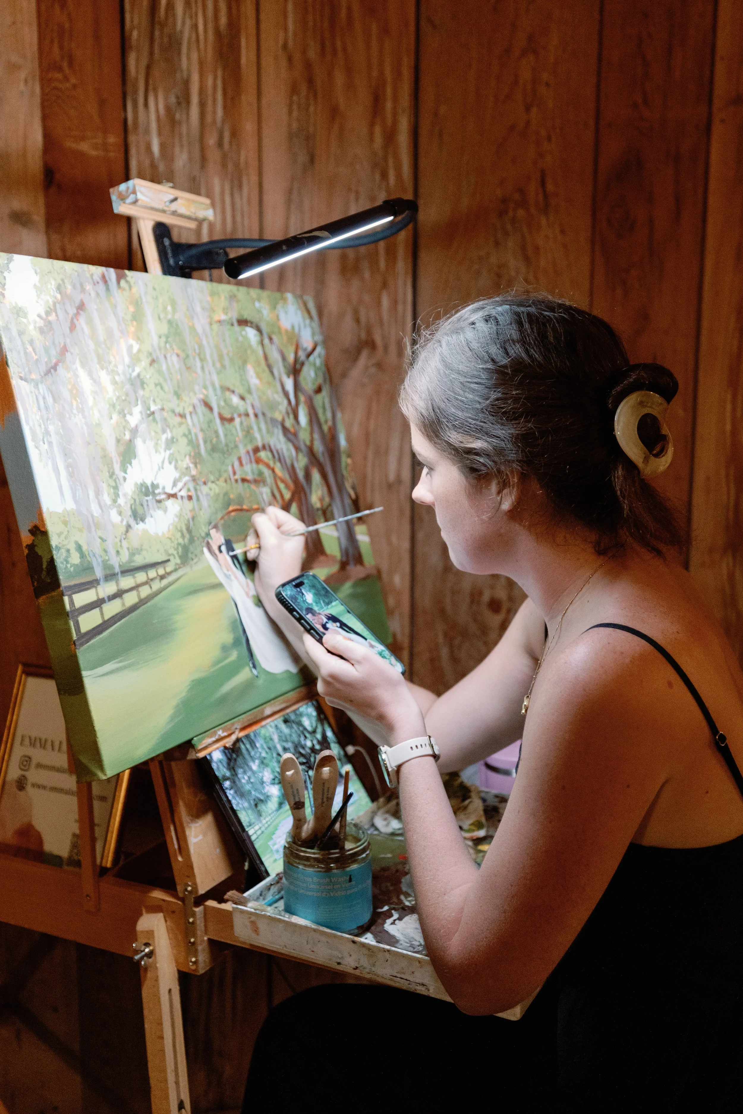 A woman painting outdoors, sitting at an easel with a landscape painting of trees and a pathway, using her phone to take a photo of her work.