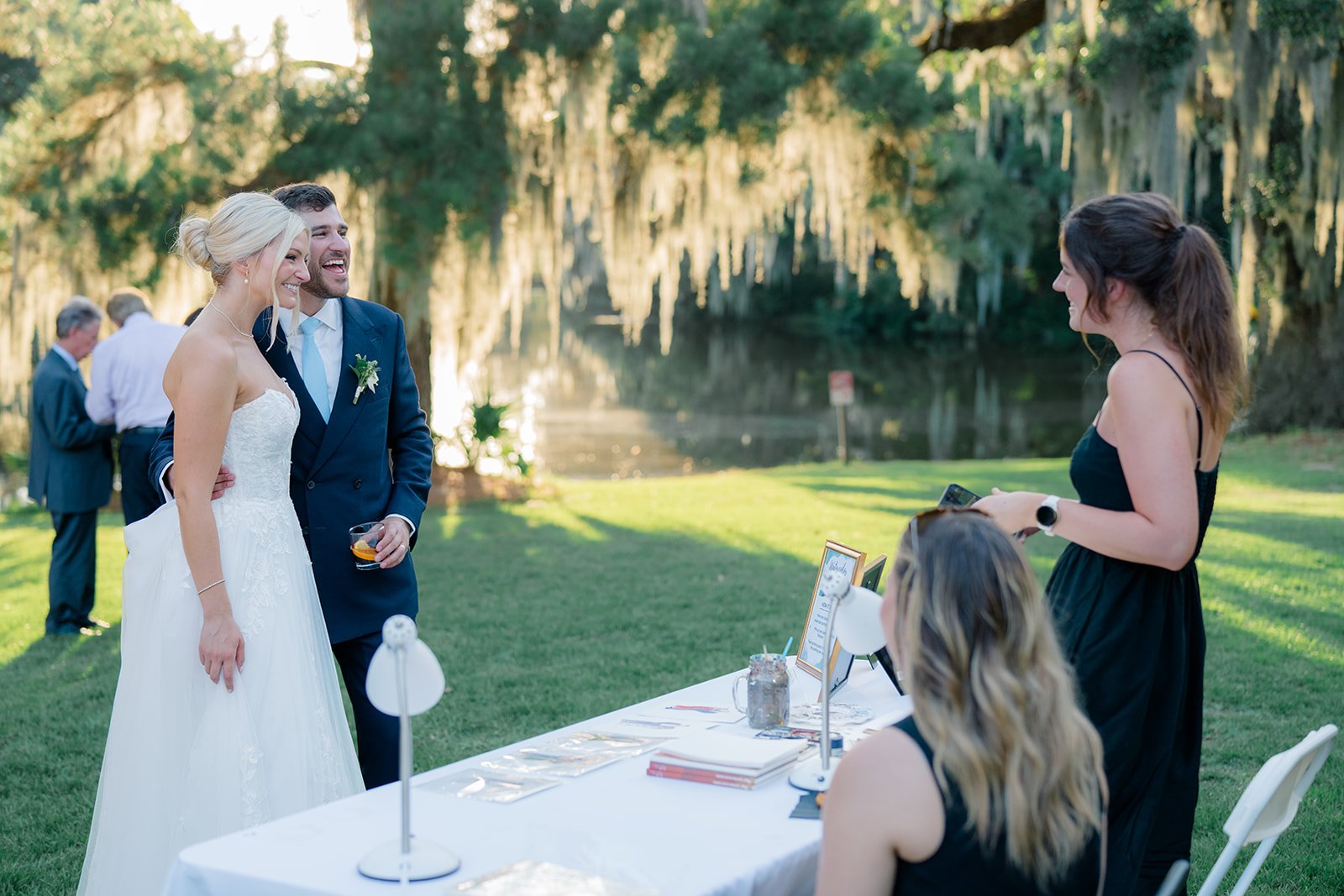 A bride and groom laugh together at their outdoor wedding reception with a woman holding a microphone and guests in the background by a pond and trees.