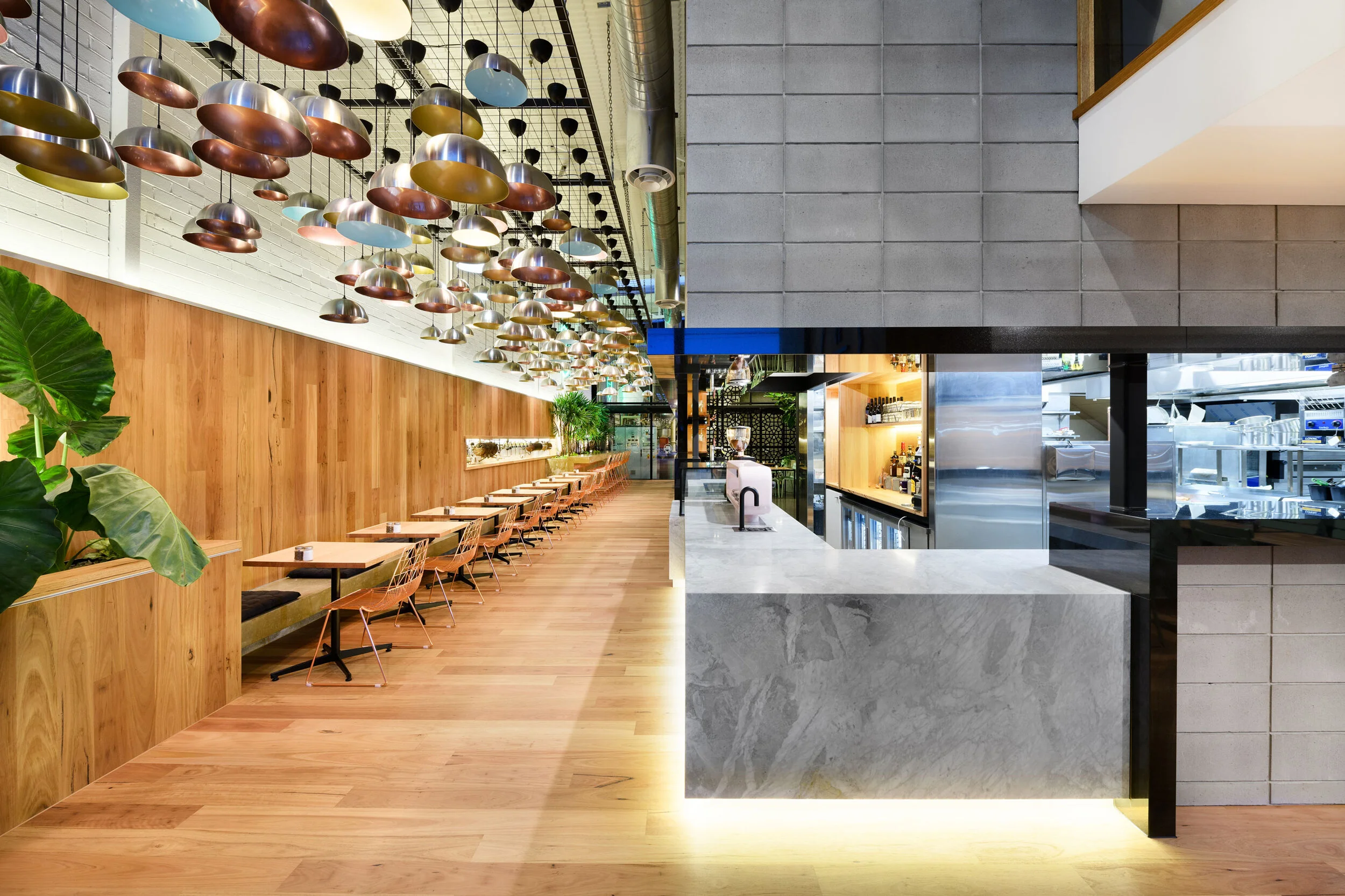 Modern restaurant interior with wooden flooring, a long row of tables and chairs on one side, a marble counter on the other, decorative pendant lights hanging from the ceiling, and a kitchen visible behind the counter.