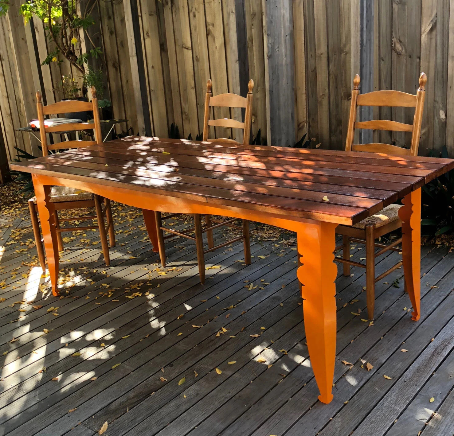 Wooden outdoor dining table with four matching chairs on a wooden deck, surrounded by a wooden fence and some leafy plants, with dappled sunlight and fallen leaves.