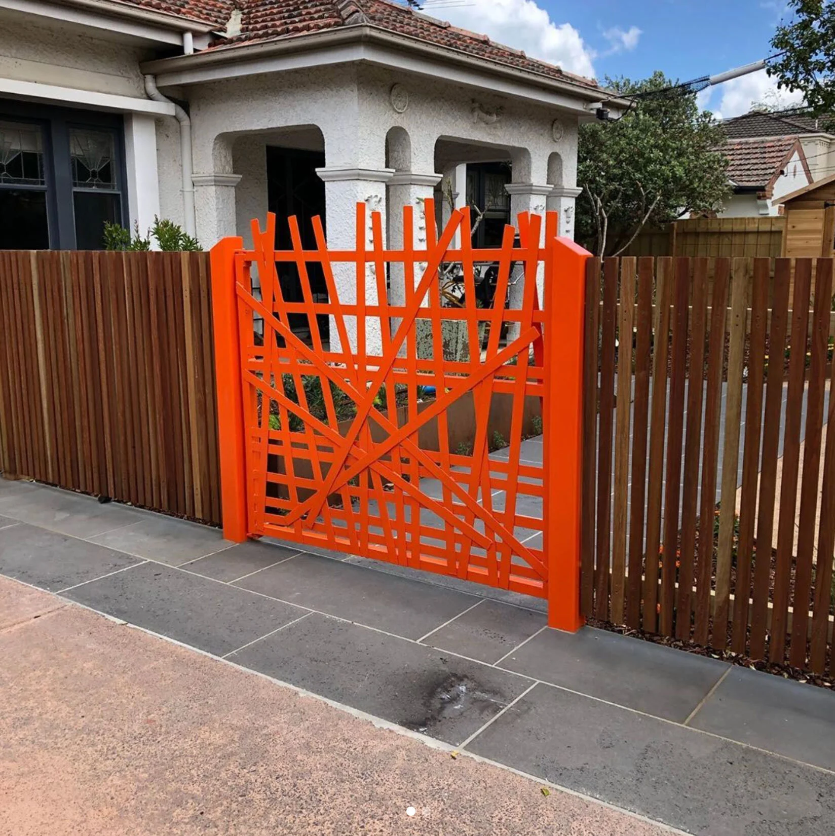 A bright orange gate with an abstract, geometric design in front of a house with a white exterior and a tiled roof. The gate is flanked by a wooden fence on both sides, and there are trees and a partly cloudy sky in the background.
