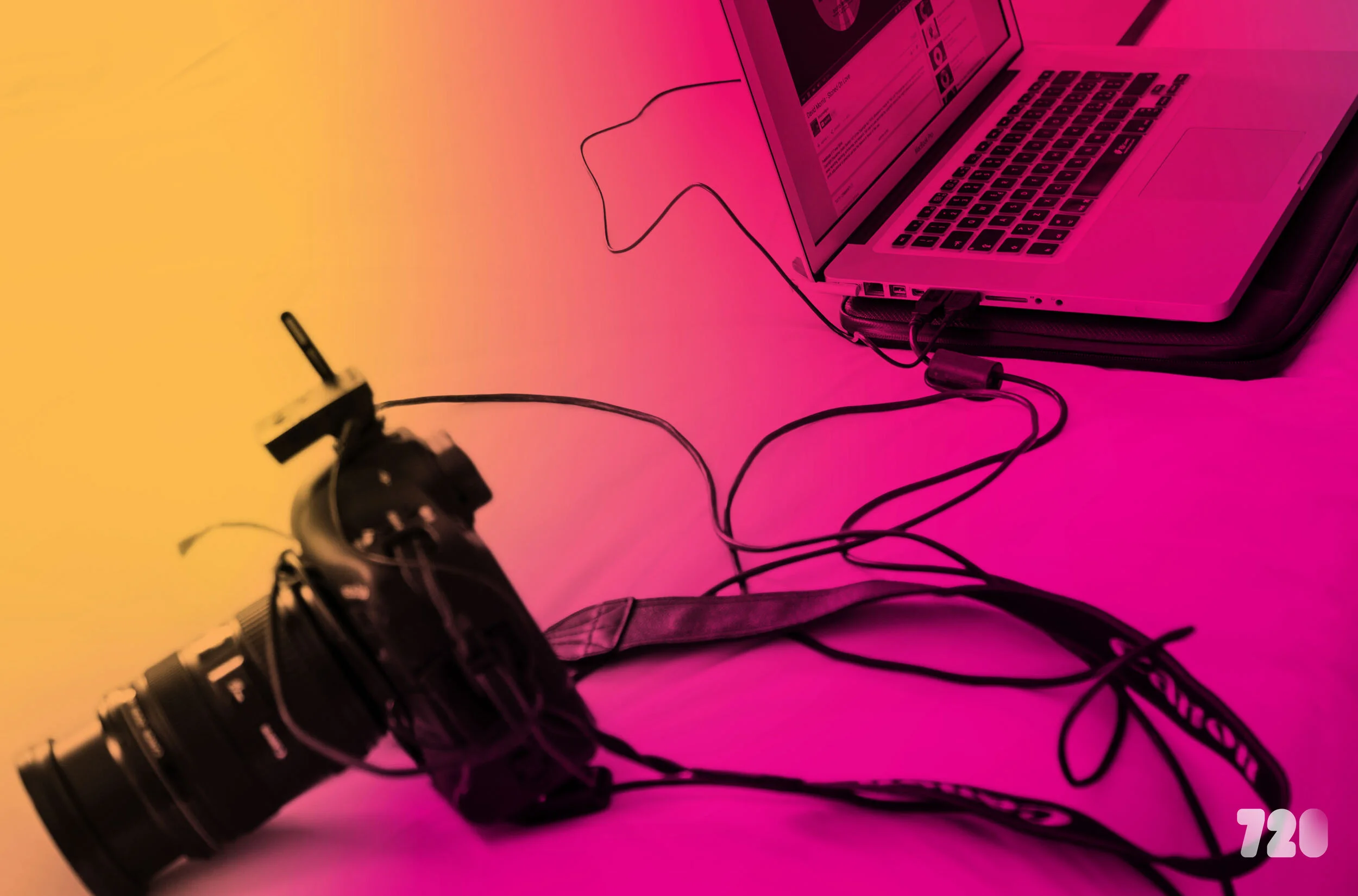 A professional camera connected with cables to a laptop, used for photography editing or shooting, on a brightly colored pink and yellow background.