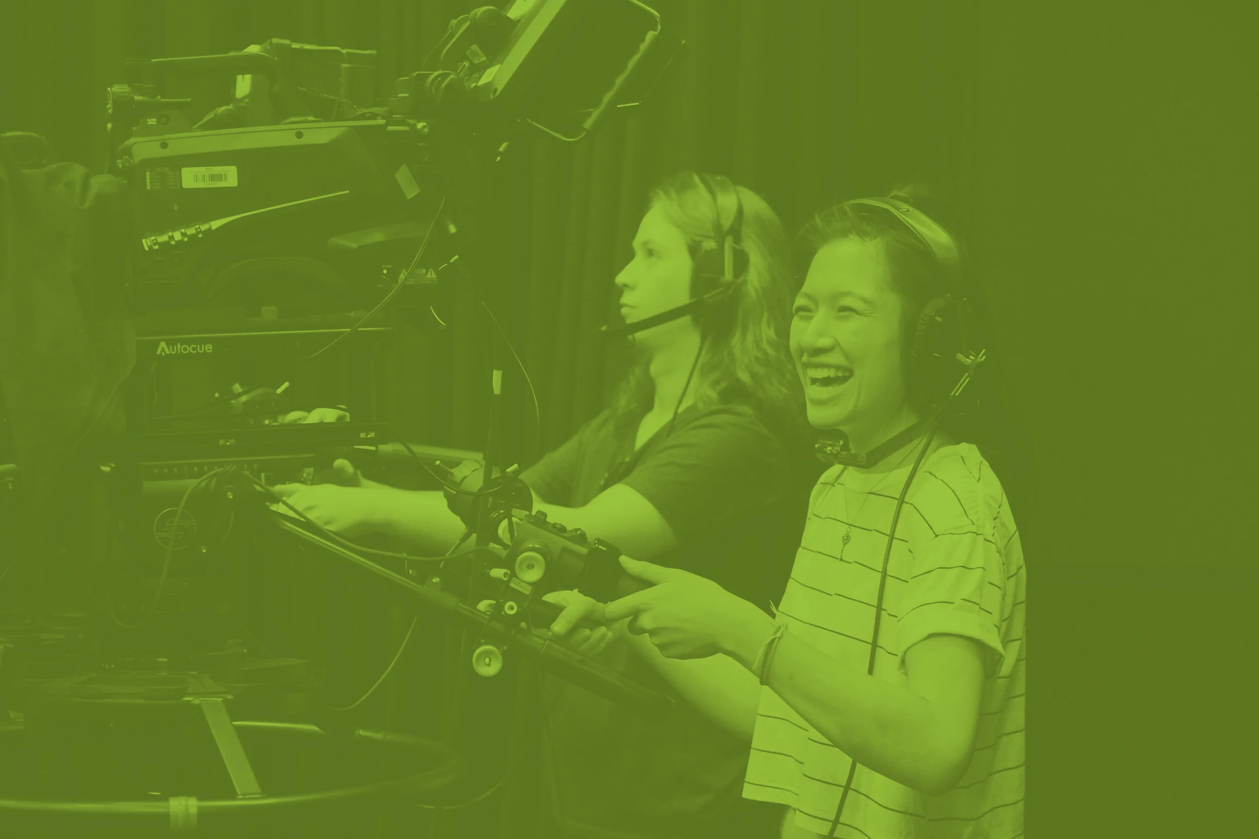 Two women wearing headsets working behind a camera on a film or television set, with one woman smiling and the other focused.