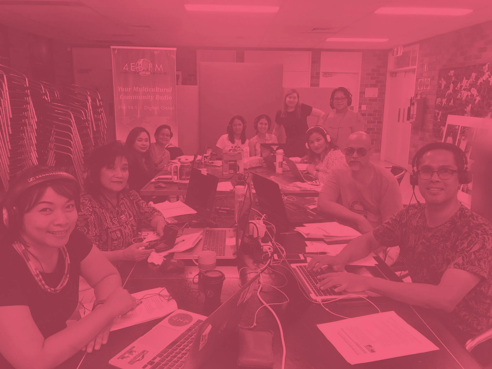 A group of people sitting around a table with laptops, microphones, and paperwork in a community radio station room. Some are wearing headphones, and there are stacked chairs and a banner in the background.