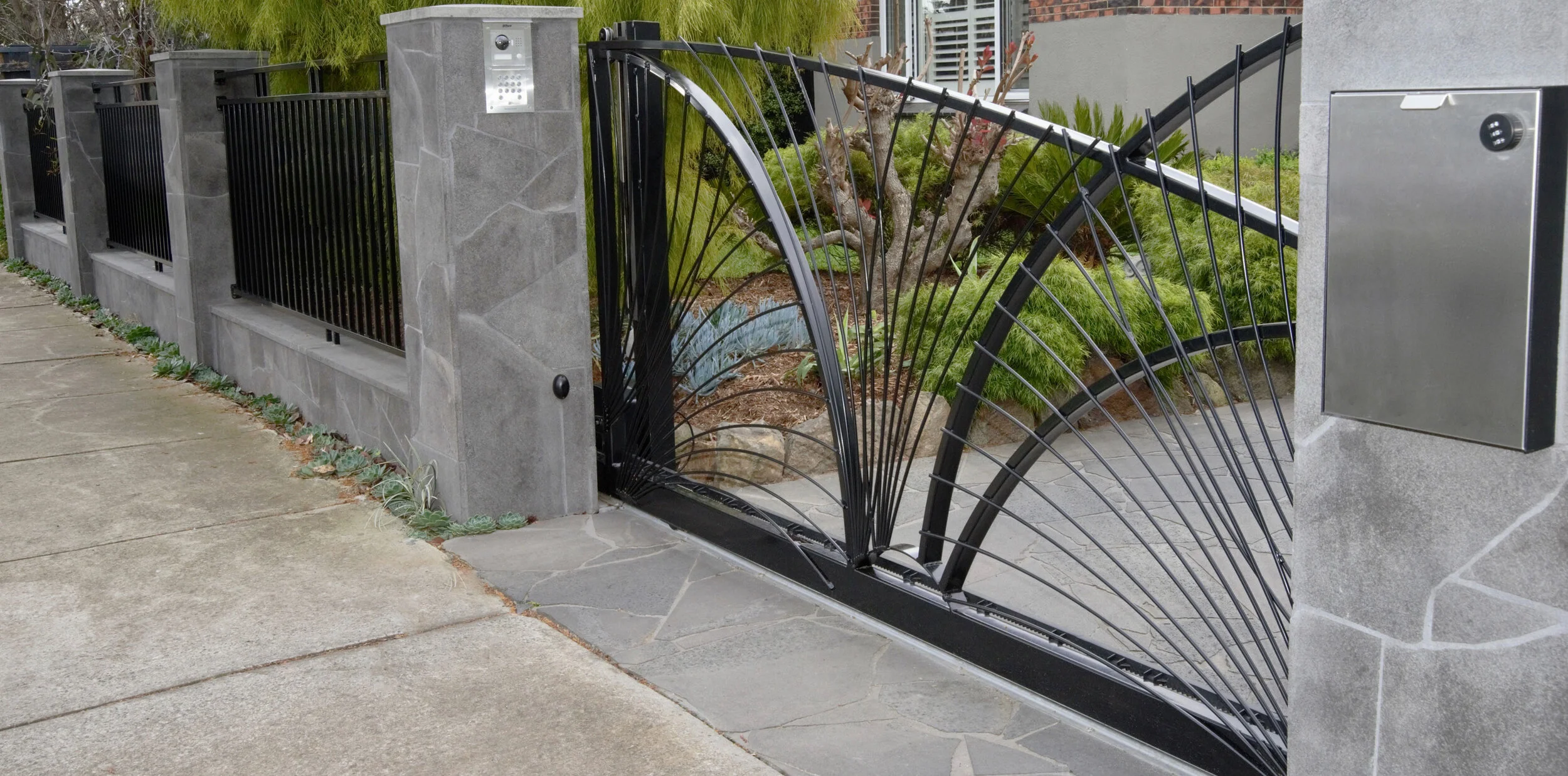 A modern exterior gate with a decorative black metal design, set between gray stone pillars that include a gate controller and mailbox. There is a sidewalk along the gate and various plants behind it.