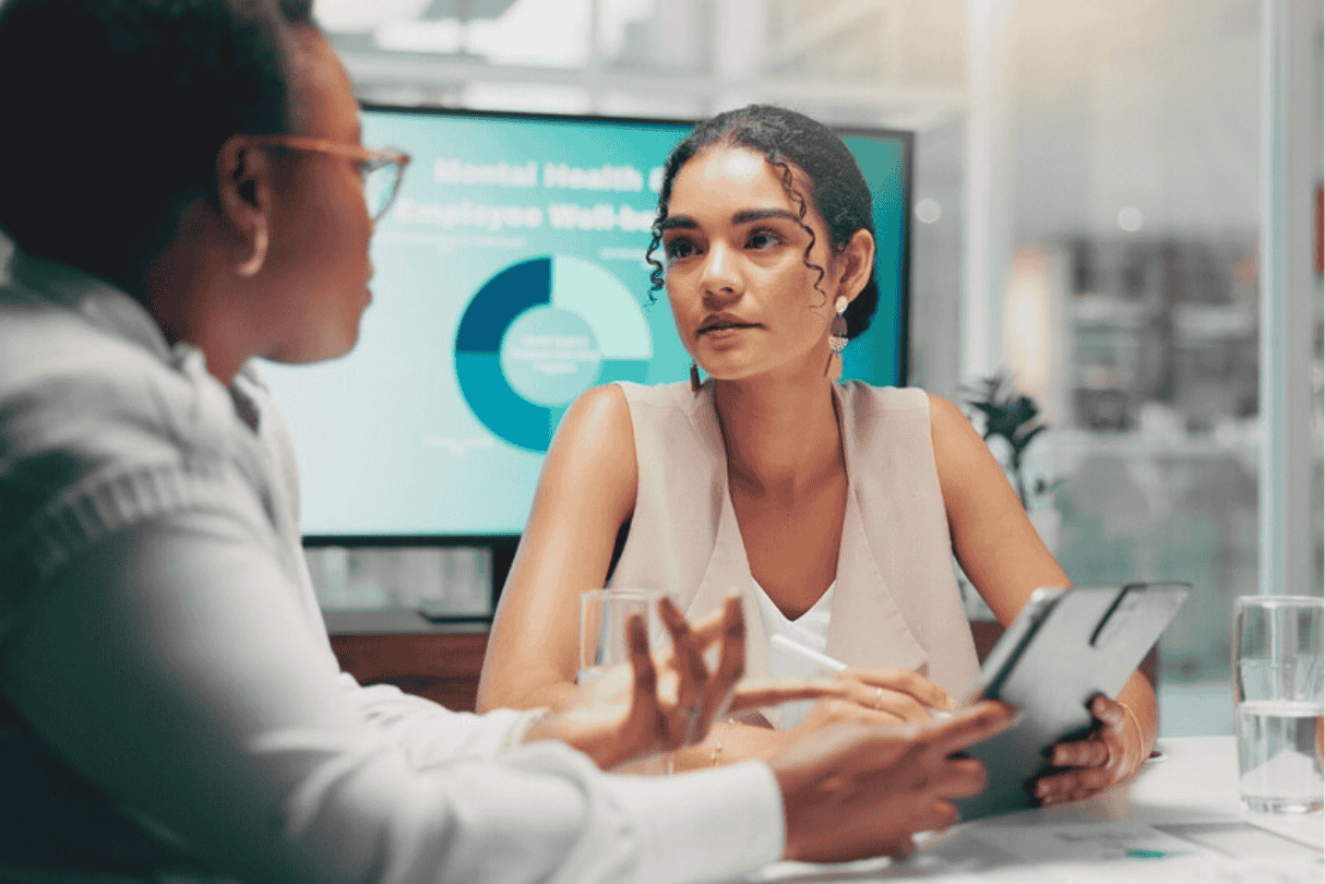 Two women in a business meeting discussing in a conference room with a digital screen displaying a pie chart in the background.