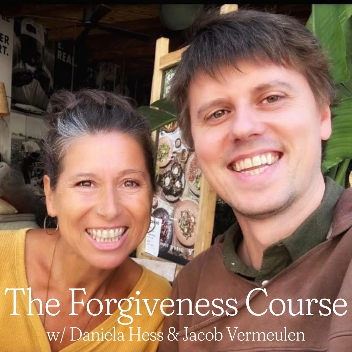 A smiling woman with dark hair in a bun wearing a yellow top, and a smiling man with light brown hair wearing a brown shirt, taking a selfie together in front of a display of artwork and decorative items in an indoor setting.