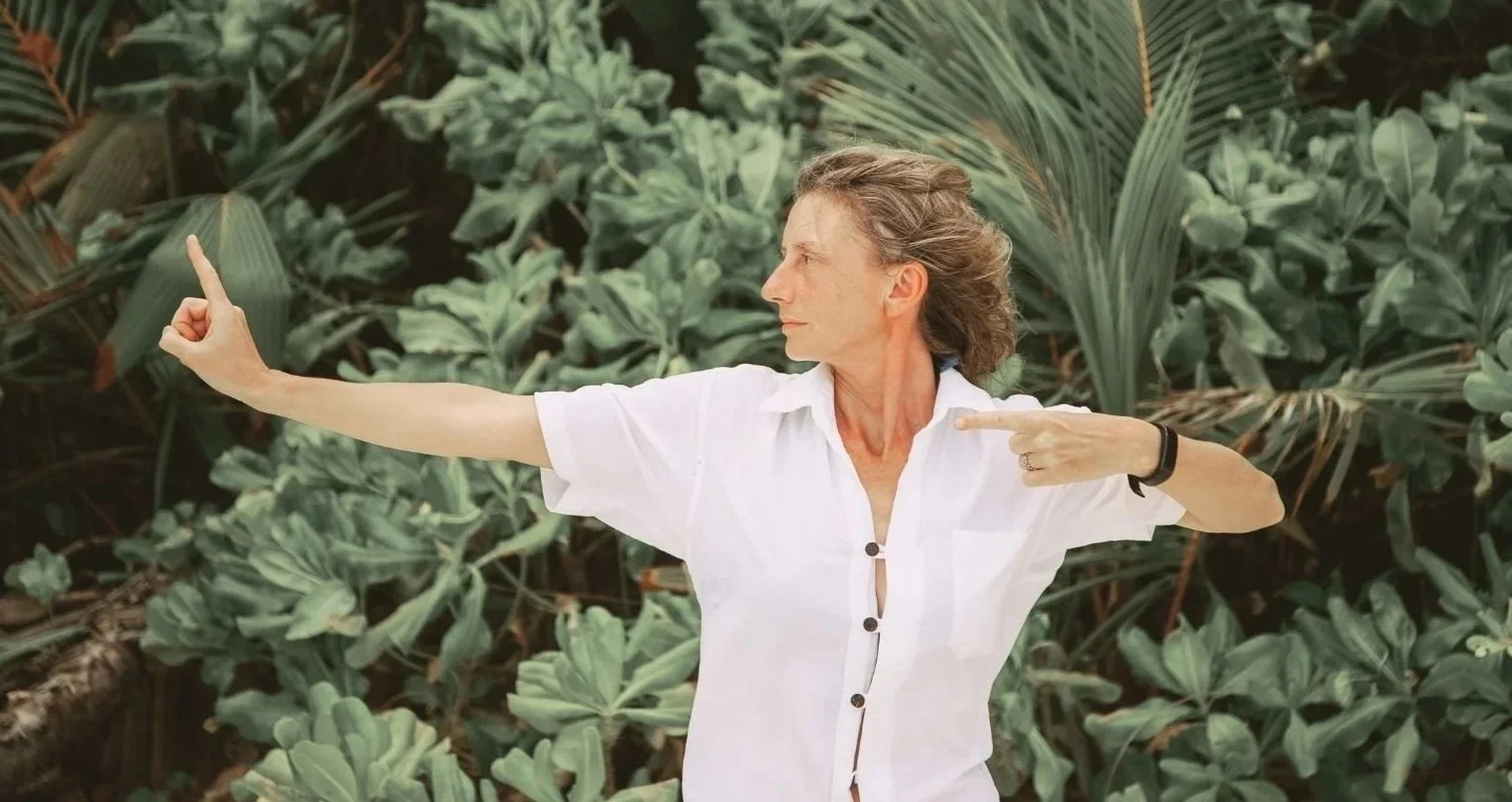 A woman in a white shirt pointing her right hand forward and her left hand toward her shoulder in a Healing Qigong stance while standing in front of dense green foliage.