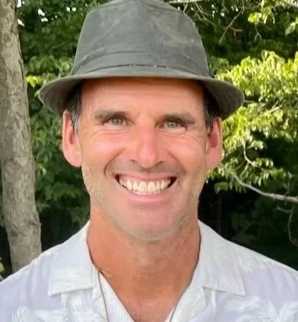 A woman with long, dark, curly hair smiling outdoors with trees in the background. Text overlay shows the name Kimberly Beekman.
