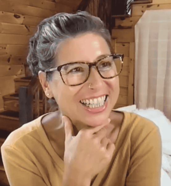 A woman named Daniela Hess with short, curly dark hair, clear glasses, and a nose ring, smiling indoors with wooden walls and a window in the background.