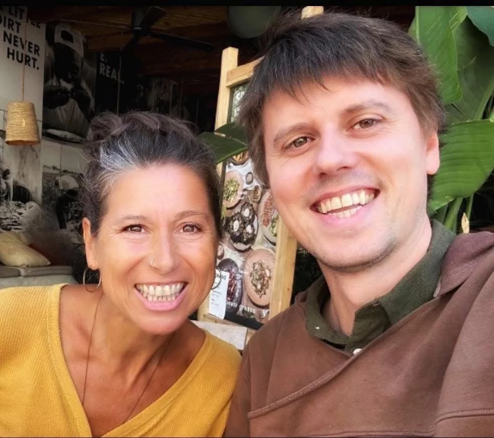 A woman with dark hair in a bun and a yellow top smiling next to a smiling man with brown hair, in a setting with green plants and decorative items in the background.