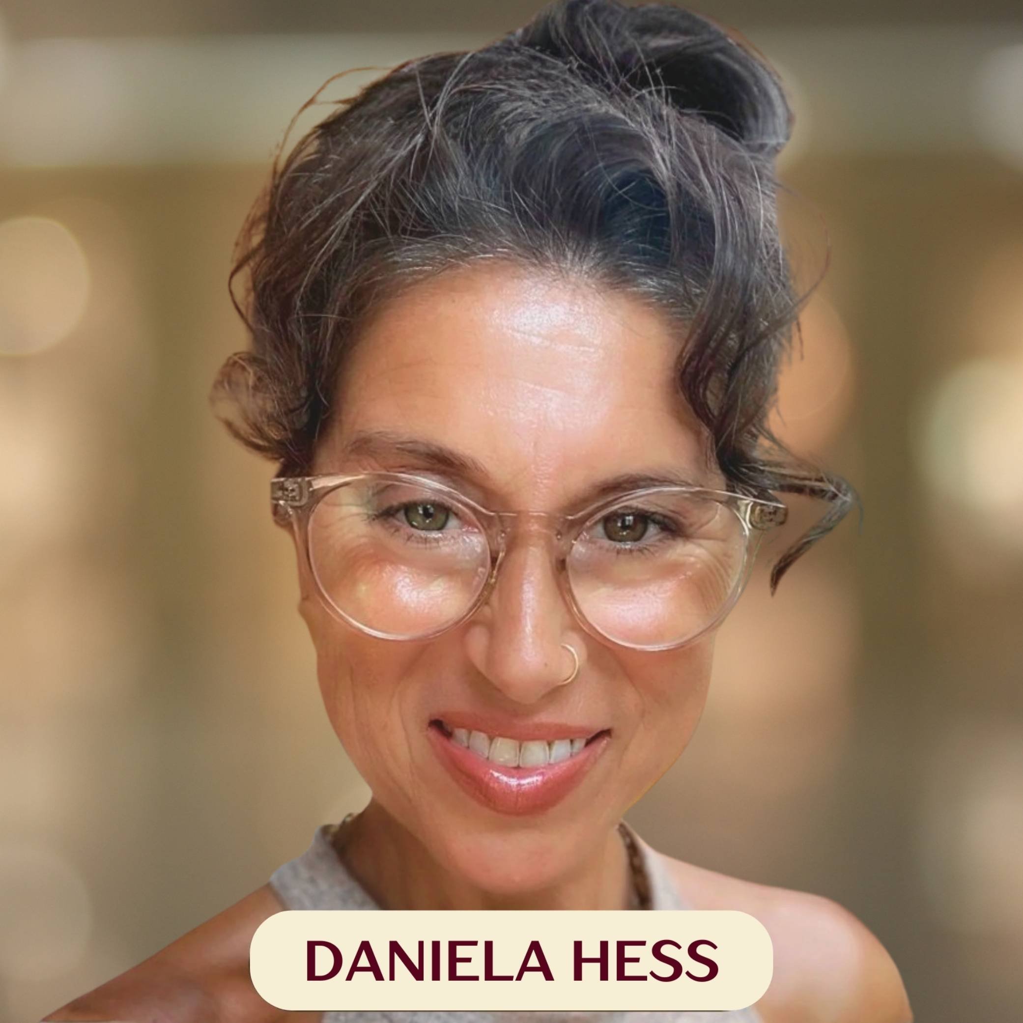 A woman named Daniela Hess with short, curly dark hair, clear glasses, and a nose ring, smiling indoors with wooden walls and a window in the background.