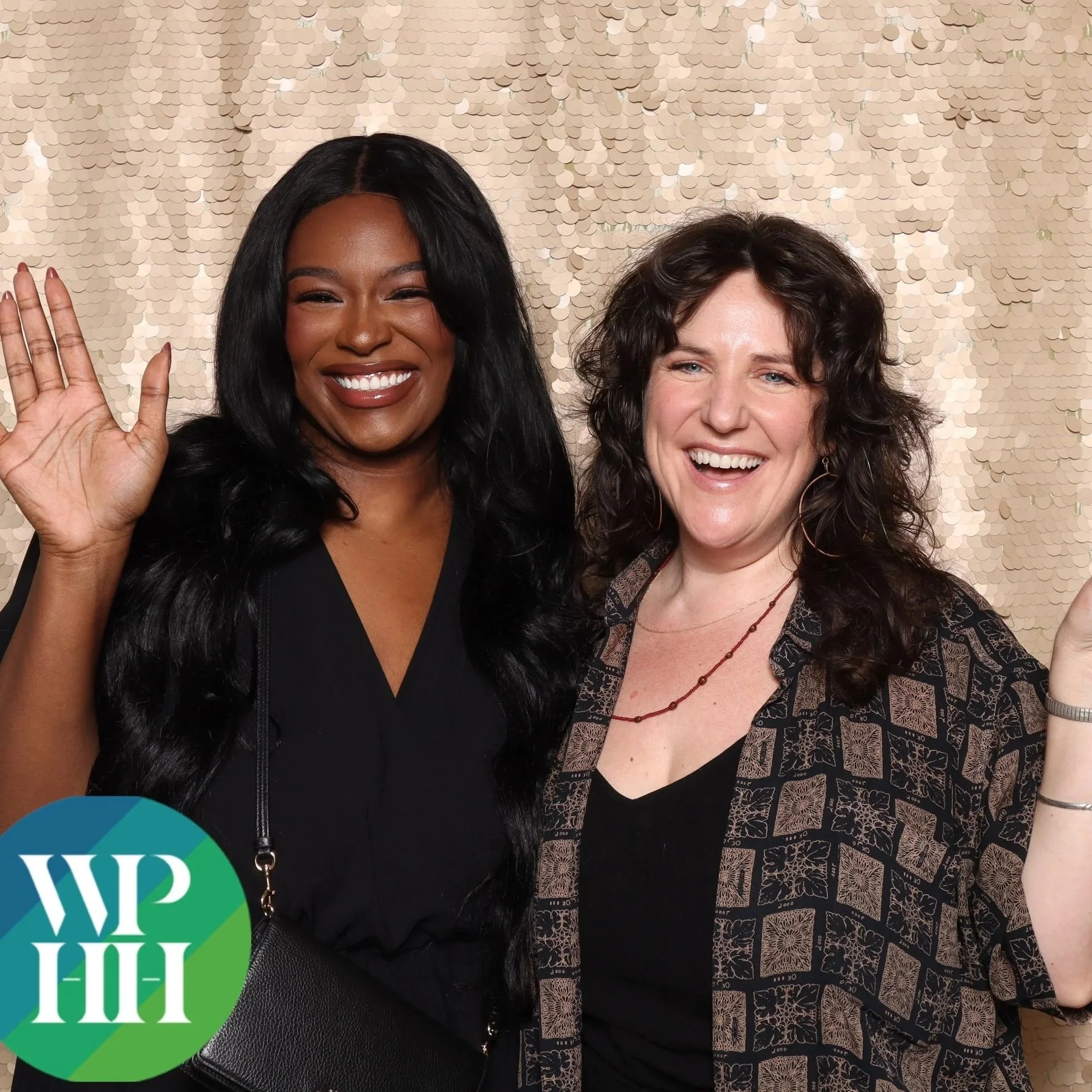 Two woman smile brightly with one hand up standing in front of a gold sequin photobooth backdrop while enjoying a wedding pro happy hour event in Portland, Oregon.