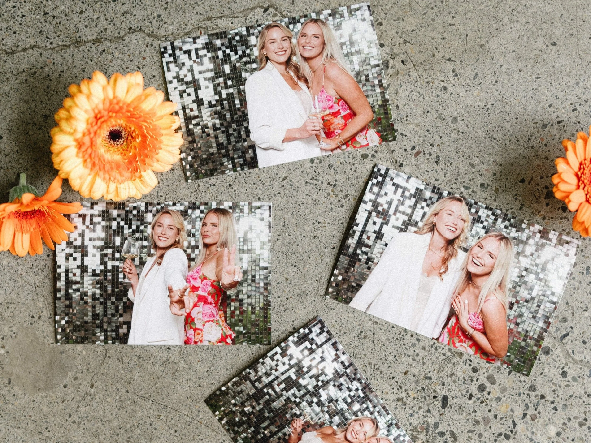 Four printed full page 4x6 photobooth photographs of two women at a celebration, laid out on concrete with orange and yellow flowers around them. Party keepsakes. Photobooth prints.