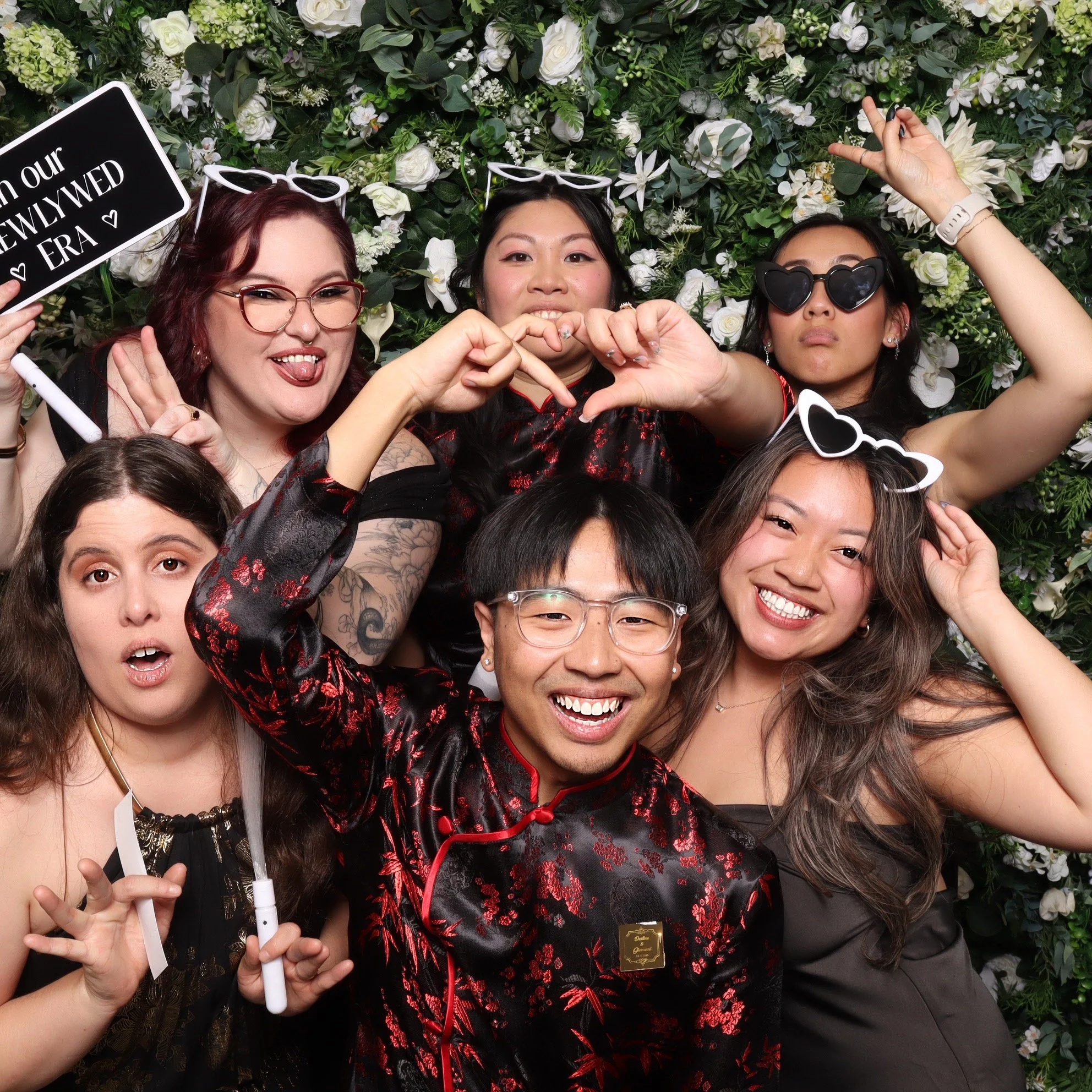 A group of six pose with black and white wedding photobooth props against a green and white flower wall backdrop at a PNW winter wedding.