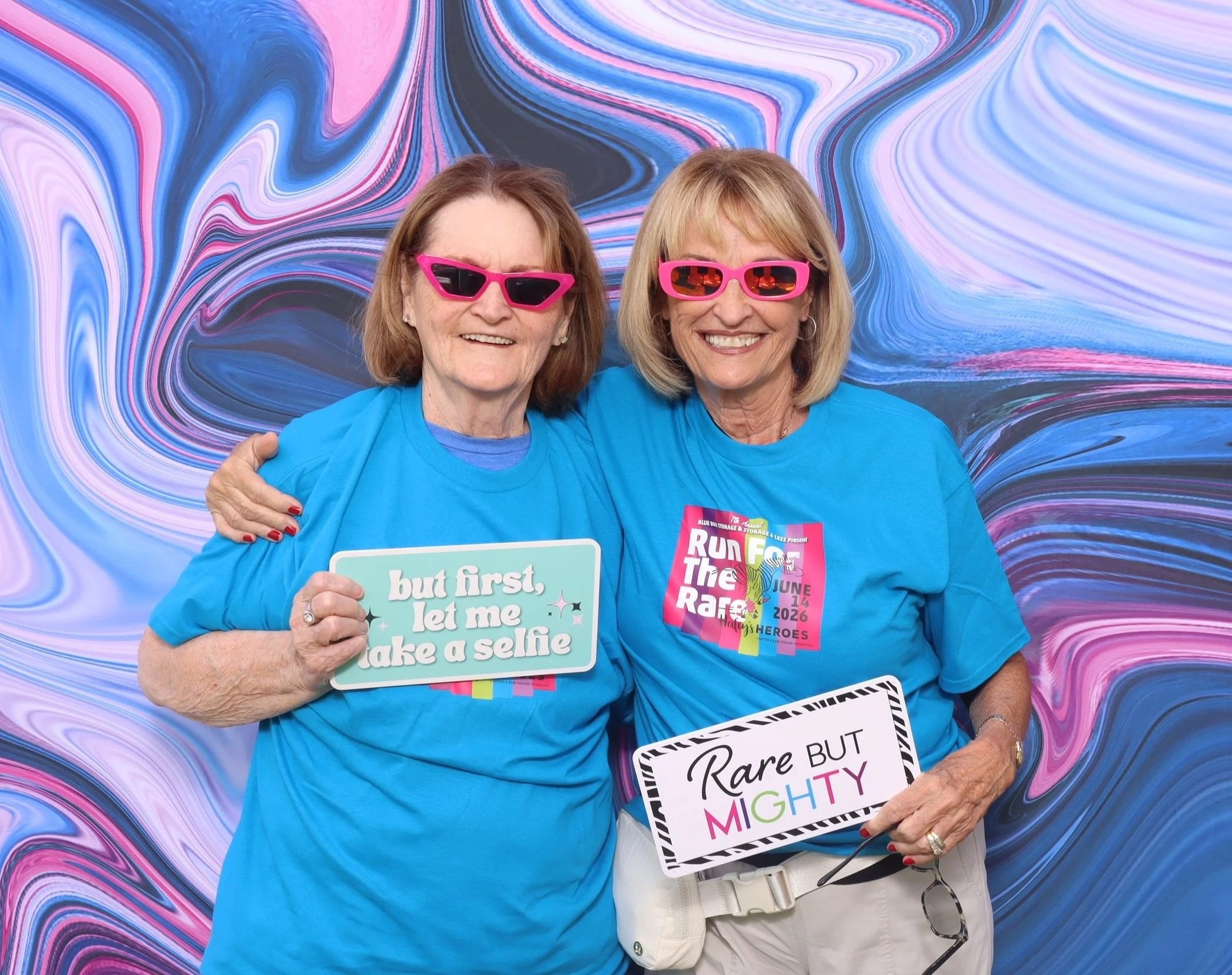 Two woman with short hair pose wearing pink sunglasses, holding brightly colored, photobooth prop signs that read "but first, let me take a selfie" and "Rare but Mighty".