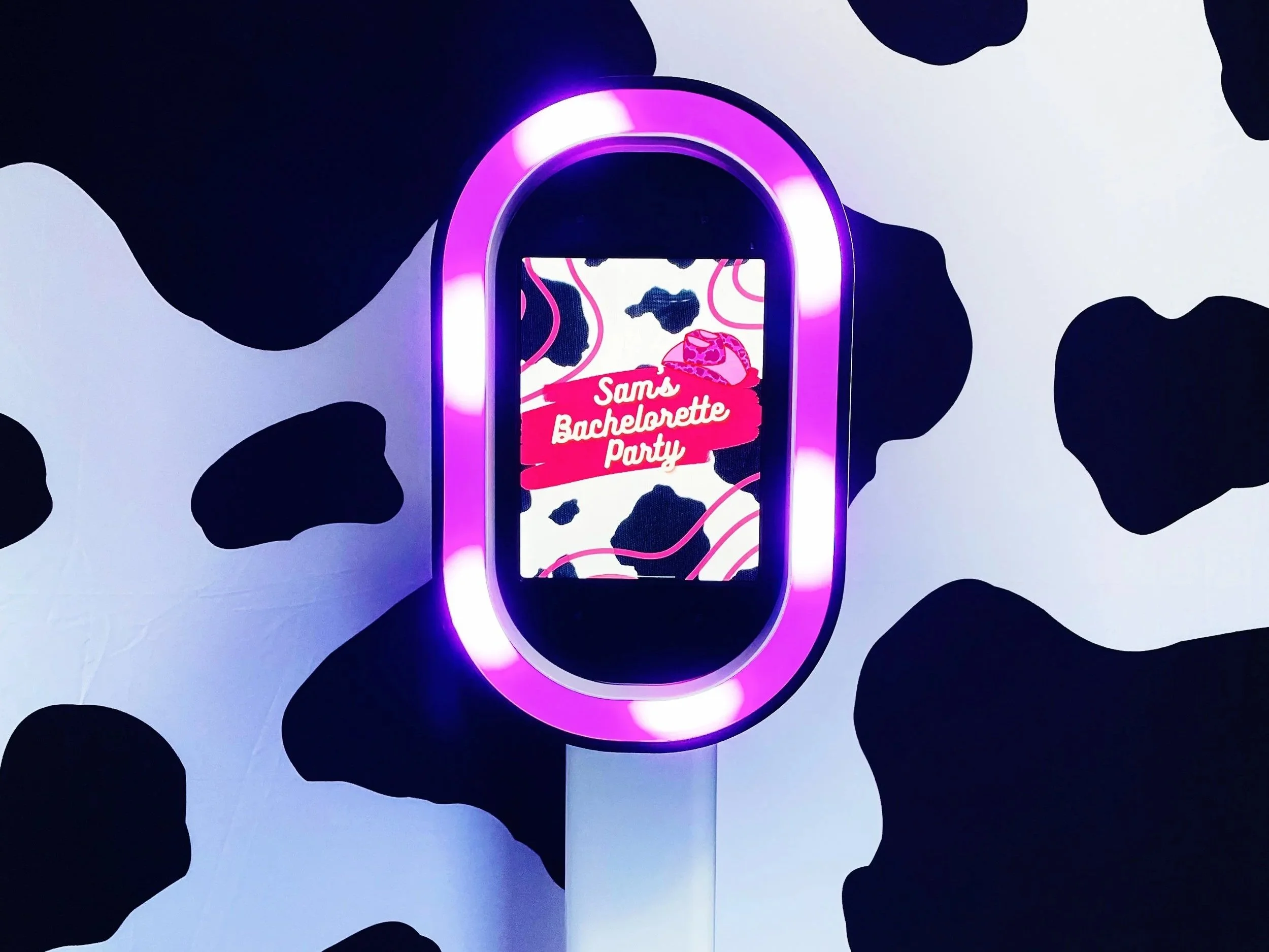 A digital photo booth with a cow print background and neon lighting, displaying a screen that reads "Sam's Bachelorette Party."