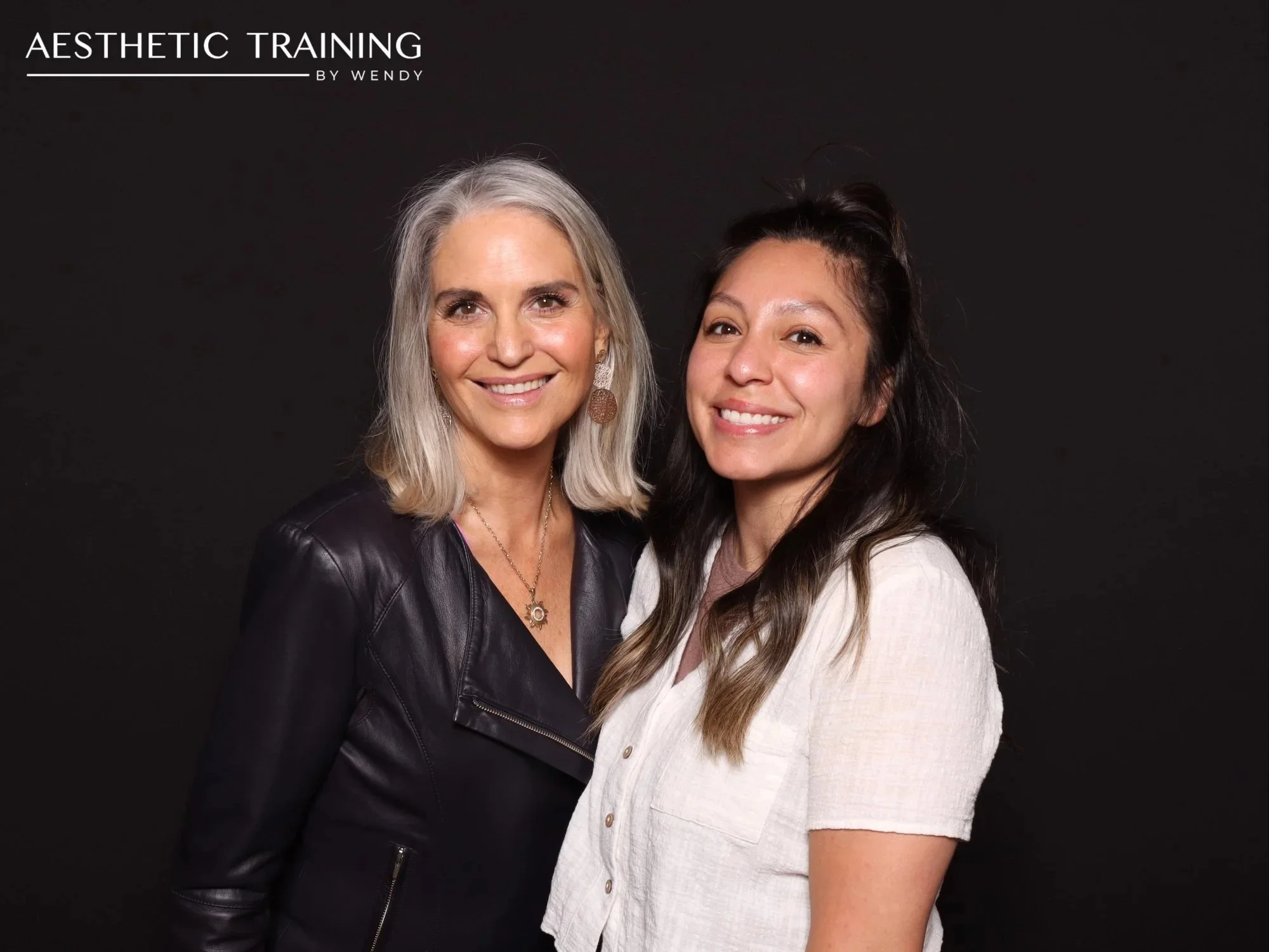 Two woman with glowing, clear skin smile in front of a black photobooth backdrop at a Aesthetic Training by Wendy event at Amaterra Winery.