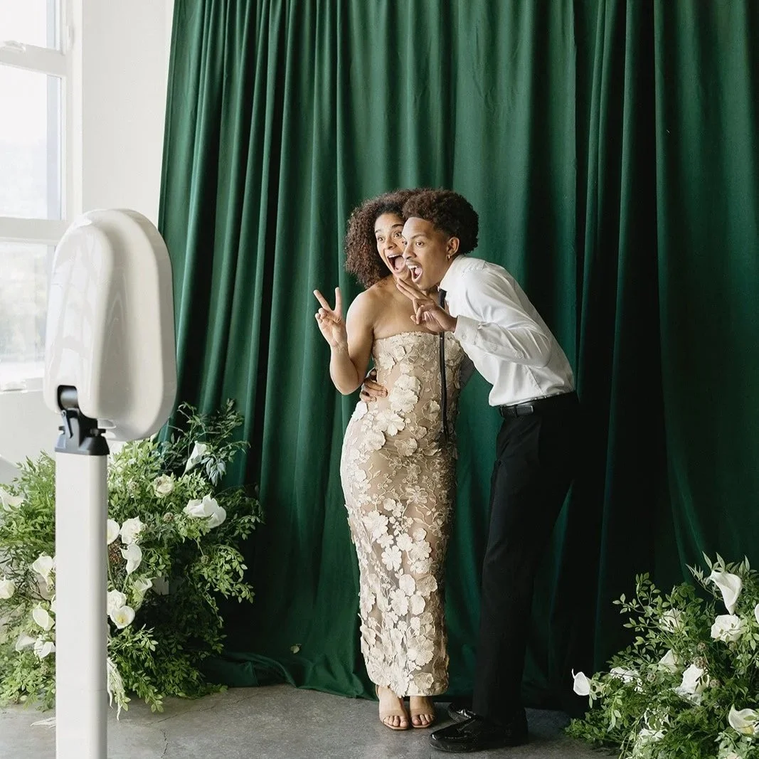 A couple poses holding up peace signs with their fingers and showing silly faces while posing infront of a green drapery photobooth backdrop at a Portland, Oregon wedding event.
