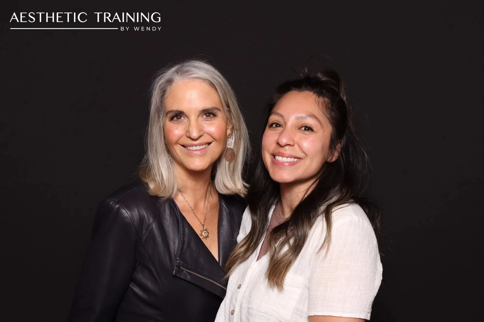 Two women smiling against a black photobooth backdrop at an event at Amaterra Winery hosted by 'Aesthetic Training by Wendy'.