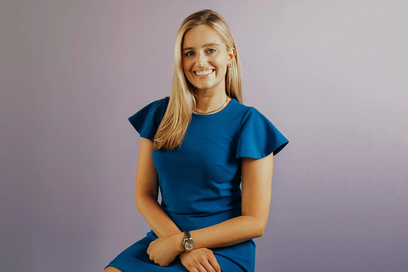 A woman with long blonde hair wearing a blue dress and watch sitting against a light purple background, smiling.