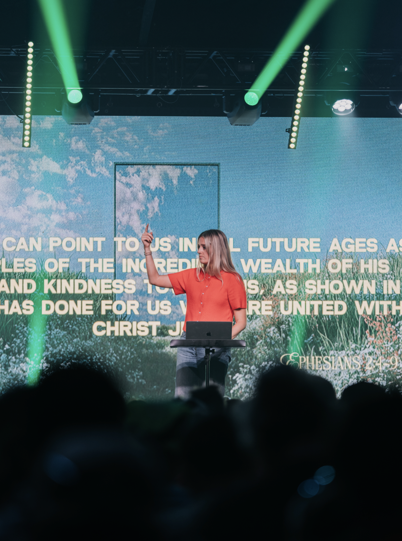 A woman in a red shirt standing behind a lectern and raising her right hand, with a large digital screen behind her displaying a quote from Ephesians 2:8-9 about salvation through faith.